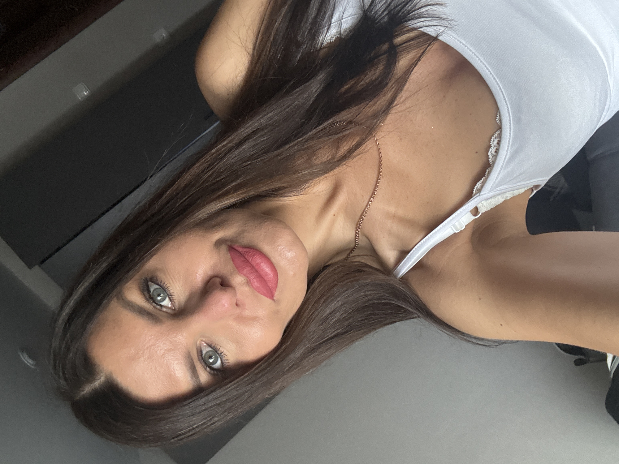 A medium close-up selfie of a woman with long, dark brown hair and light green eyes is shown. She is wearing a white tank top with lace trim. Her lips are painted a vibrant pink. She has a delicate gold chain necklace on. The background is out of focus, appearing to be an indoor setting with gray walls and dark furniture. There is no identifiable information about the location in the city of Chaikovsky, Russia.