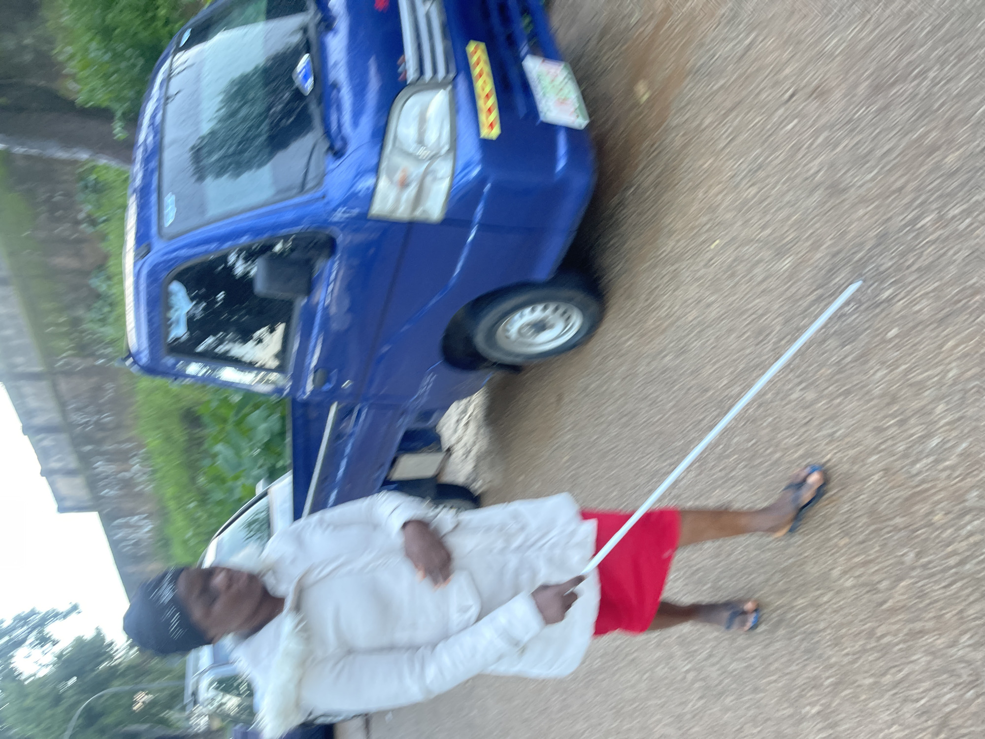 A woman, appearing to be visually impaired and using a white cane, walks on a paved road in Rafin Par, Nigeria. She is wearing a white coat with a fur-lined hood and a red skirt. Her cane is extended in front of her as she walks. A blue pickup truck is parked to her right, and a white car is visible behind her. The environment appears to be an outdoor urban setting, with some greenery and a wall in the background. The lighting suggests it is either early morning or late afternoon. The image is slightly motion blurred, indicating movement.