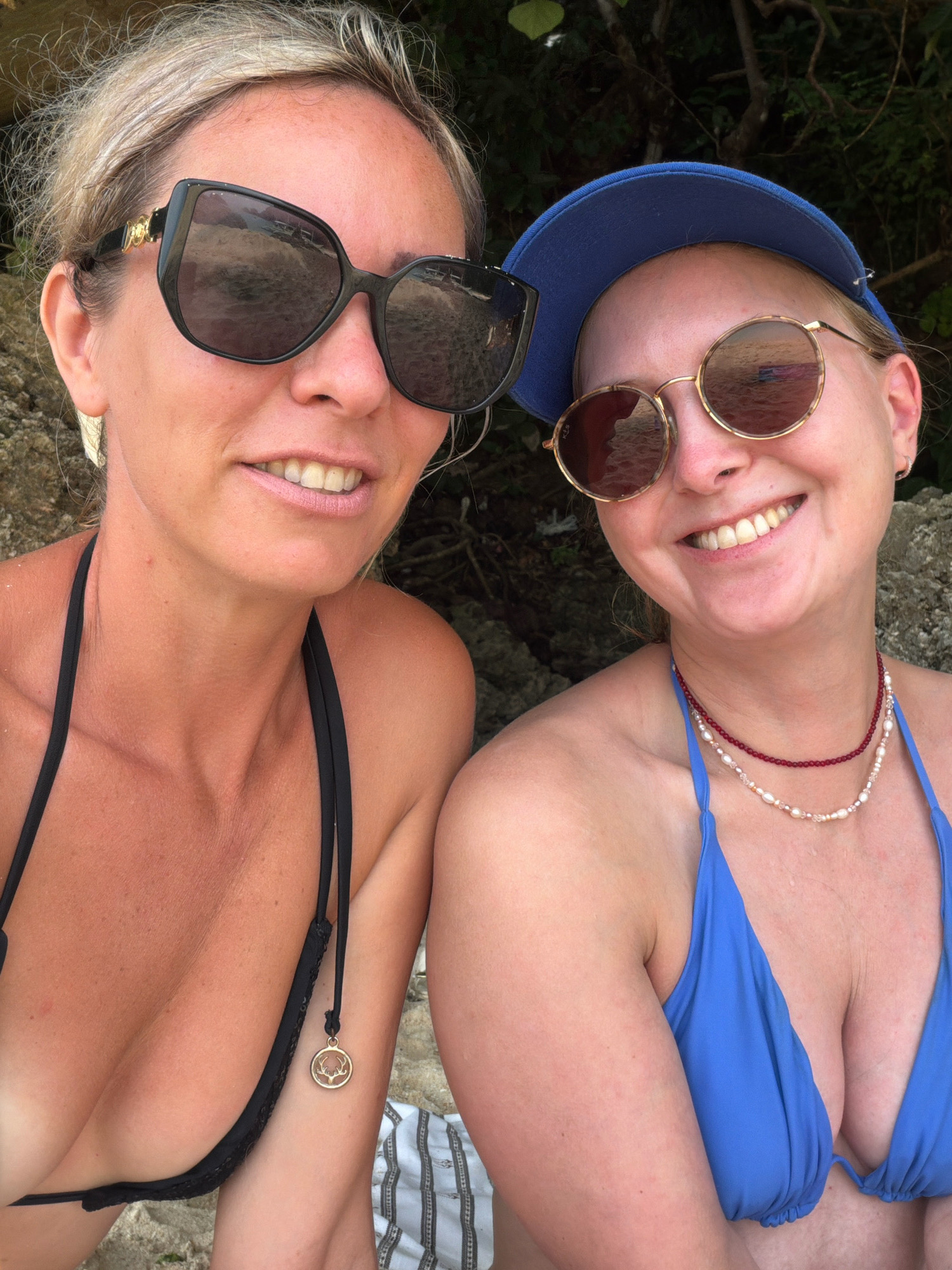 Two women are photographed from the chest up, posing for a selfie in an outdoor environment. The woman on the left has blonde hair pulled back and is wearing large, dark sunglasses and a black bikini. She is smiling with her teeth showing. The woman on the right is wearing a blue baseball cap, round sunglasses, and a blue bikini. She is also smiling broadly. Both women have fair skin and appear to be on a beach, with sand and rocks visible in the background. The vegetation suggests a tropical location, consistent with Malay, Philippines, which is known for its beaches and islands.
