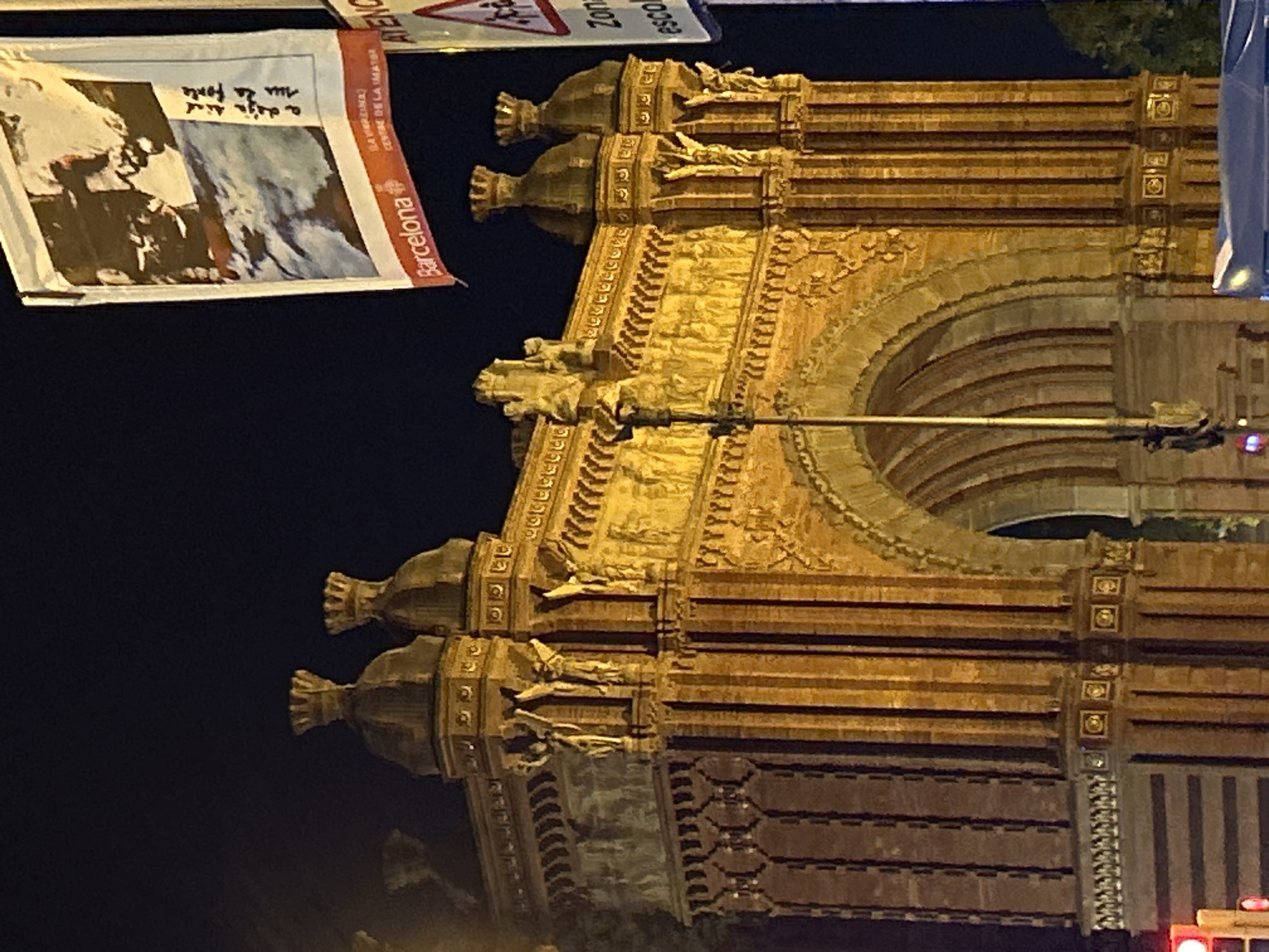 The image depicts the Arc de Triomf in Barcelona, Spain, at night. The arch is illuminated and shows its detailed sculptural elements. To the left of the arch is a poster with an image and the word "Barcelona" on its right side. A street sign with what looks like a triangle is partly visible as well. The night sky is dark and clear.