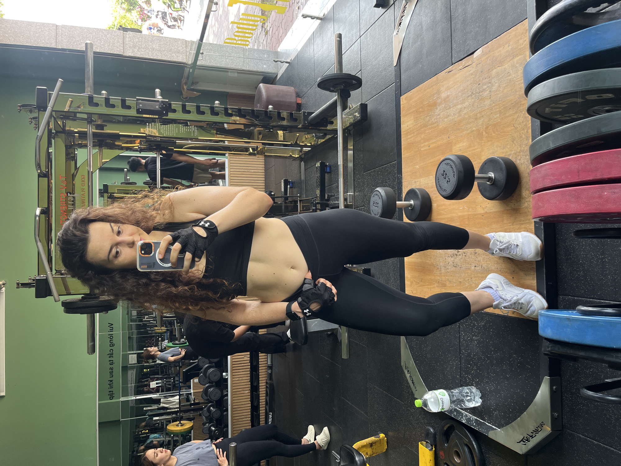 The image depicts a woman with curly brown hair taking a selfie in a gym. She is wearing a black sports bra, black leggings, black workout gloves, and white athletic shoes. Her abdomen is exposed, revealing toned muscles. In the background, other gym equipment such as weight racks, dumbbells, and barbells are visible. A water bottle is placed on the floor near her feet. The gym appears to be indoors, with green walls and natural light coming from a large window. Outside the window, a street scene with motorcycles and people can be observed, suggesting an urban environment. The text on the wall in the reflection of the mirror reads "Bodybuilding" and the location is identified as Ham Thang Ward, Vietnam. The overall scene captures a moment of a fitness workout in a public gym.