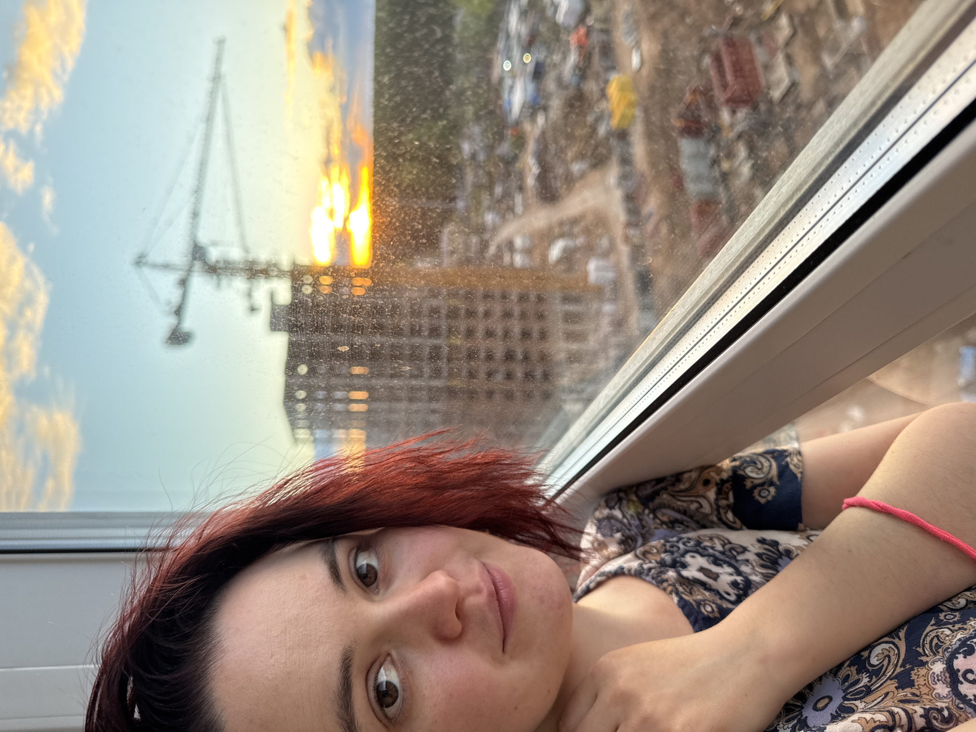 A woman with auburn hair looks at the camera in front of a window. Behind her is a construction site, a building in progress, and a crane. The sunset is visible above the construction site. The woman wears a patterned dress and a pink bracelet. The window frame is white. Based on the architectural style and visible landmarks, the location is likely Izhevsk, Russia.