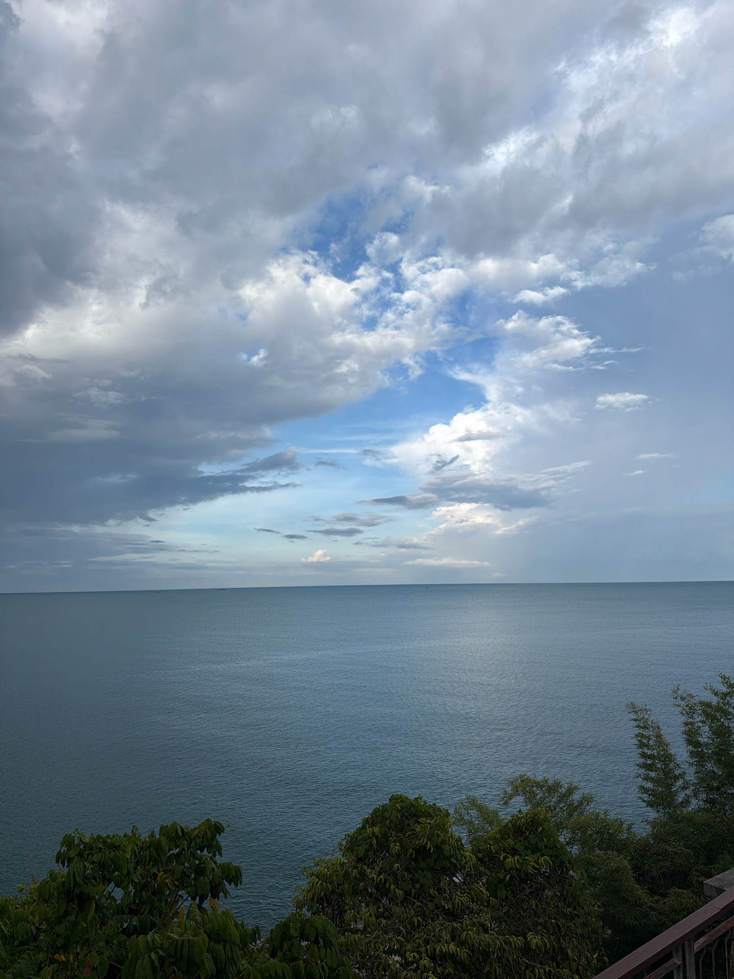 The image depicts a serene seascape with a cloudy sky over a calm ocean. The foreground features lush green foliage and a dark brown railing, suggesting a vantage point from a balcony or elevated area. The water is a deep blue-gray, reflecting the overcast sky. The horizon is clearly defined, separating the sea from the sky. The sky is filled with a mix of dark, heavy clouds and patches of brighter blue sky, indicating a dynamic weather pattern. A few faint, dark specks on the water's surface might be distant boats. The location is identified as Ko Samui District, Thailand, a tropical island known for its beaches and natural beauty.