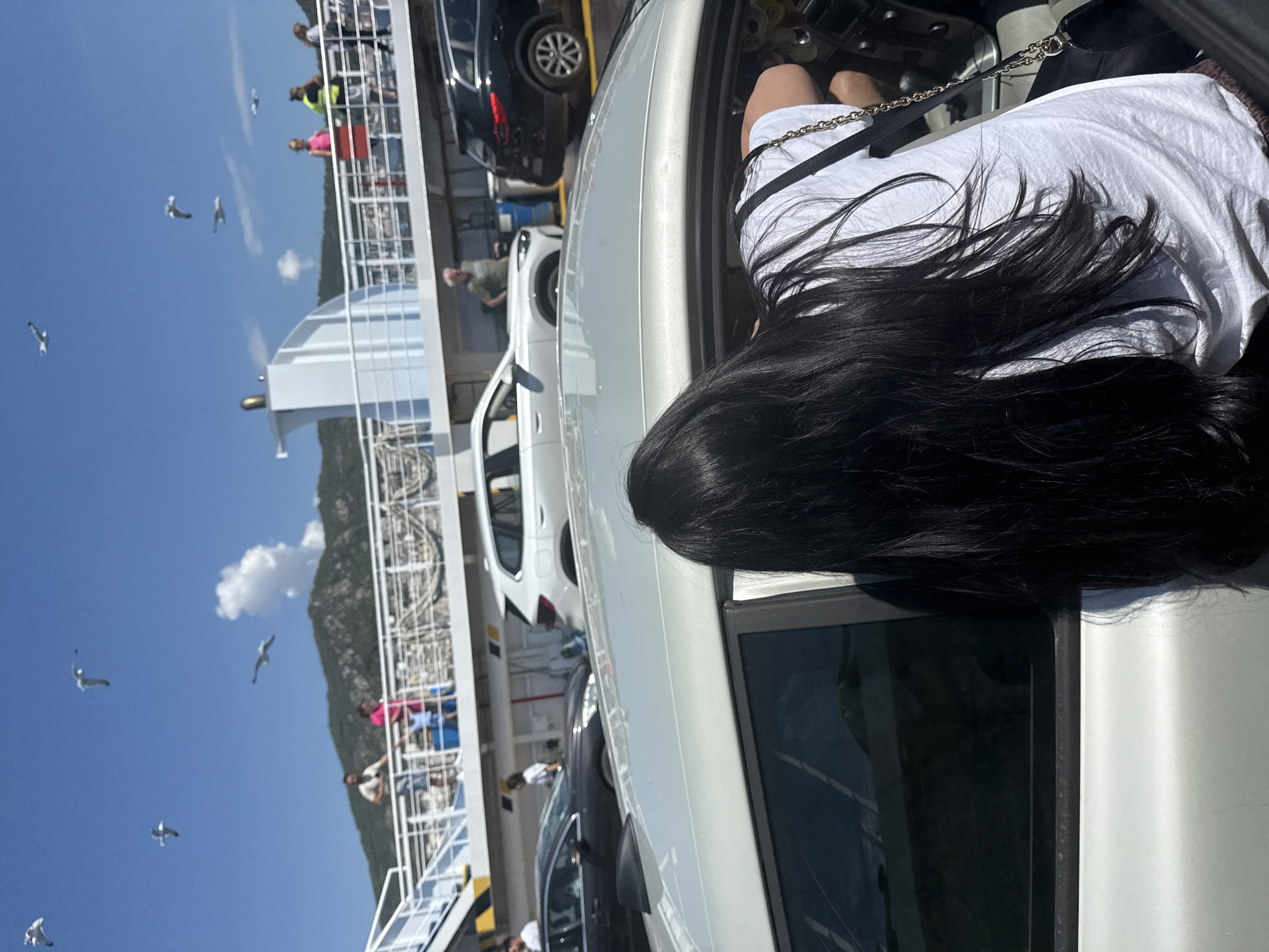The image depicts a ferry deck with several cars parked in rows, suggesting transport across water. In the foreground, a woman with long black hair is visible from behind, leaning into the driver's side of a silver car. She is wearing a white t-shirt and a dark purse with a chain strap is slung over her shoulder. Other cars, including a white SUV and a black SUV, are parked behind her. In the background, people are standing on an upper deck of the ferry, observing the surroundings. Seagulls are seen flying in the clear blue sky above. The ferry appears to be moving, as indicated by the presence of the open water and the distant, mountainous coastline, which is likely the coast of Kavala, Greece, given the context. The scene captures a moment of travel and observation on a sunny day.
