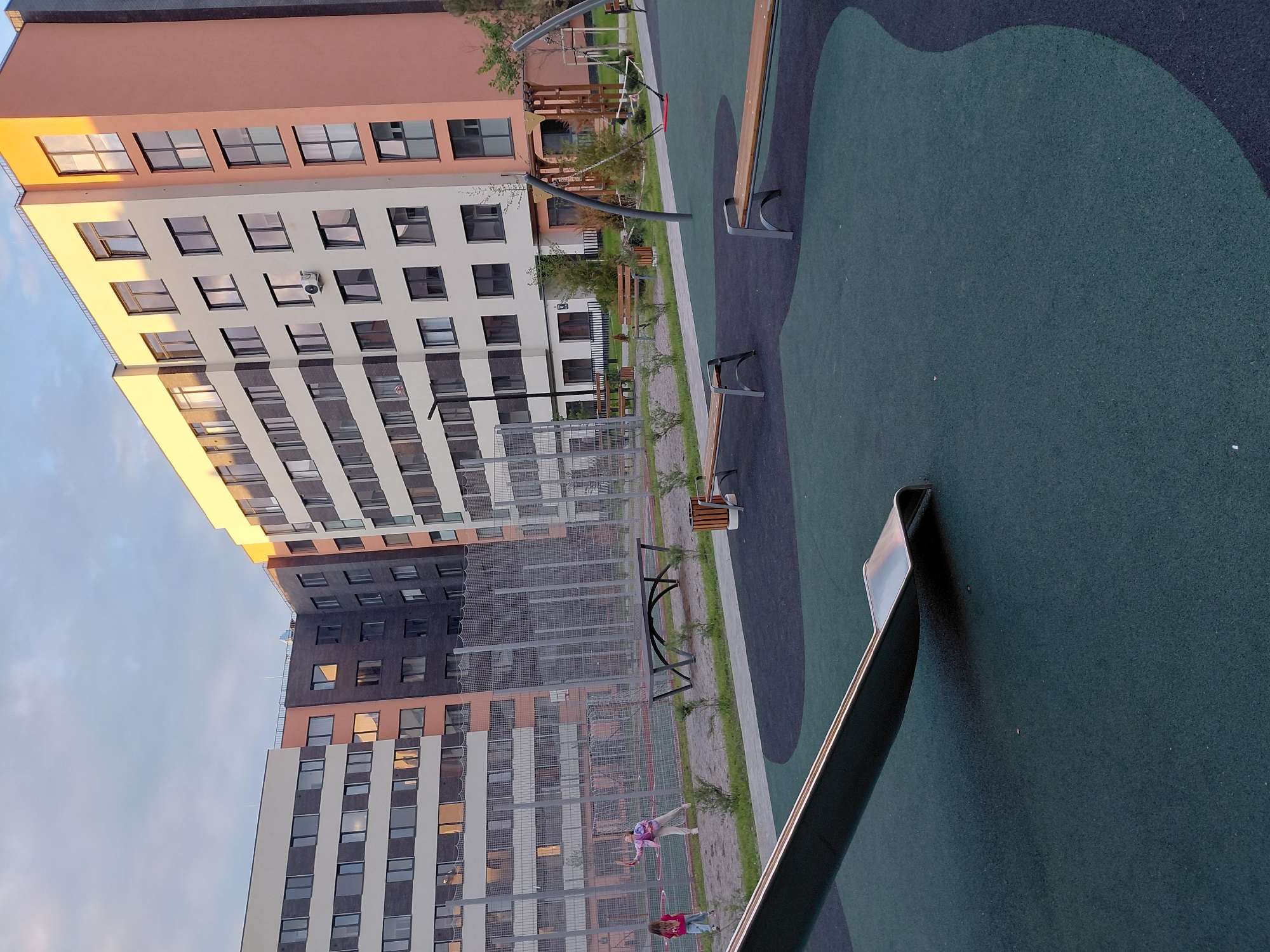 This is an outdoor photograph of a modern apartment complex playground in Guryevsk, Russia. In the foreground, a portion of a silver slide curves into a dark green rubberized surface. To the right, two dark green benches with silver metal legs are positioned on a similar surface, bordered by a lighter gray path. In the middle ground, two young girls are playing badminton on a sandy court surrounded by a chain-link fence. One girl, wearing a pink jacket and white pants, is in mid-swing. The other girl, wearing a pink shirt and blue jeans, stands nearby. Beyond the court is a long, dark gray bench with a curved design. In the background, a tall, multi-story apartment building with a beige and orange facade features numerous windows. The building is partially illuminated by the setting sun. The sky above is a mix of blue and white clouds. The scene depicts a recreational area within an urban residential environment.