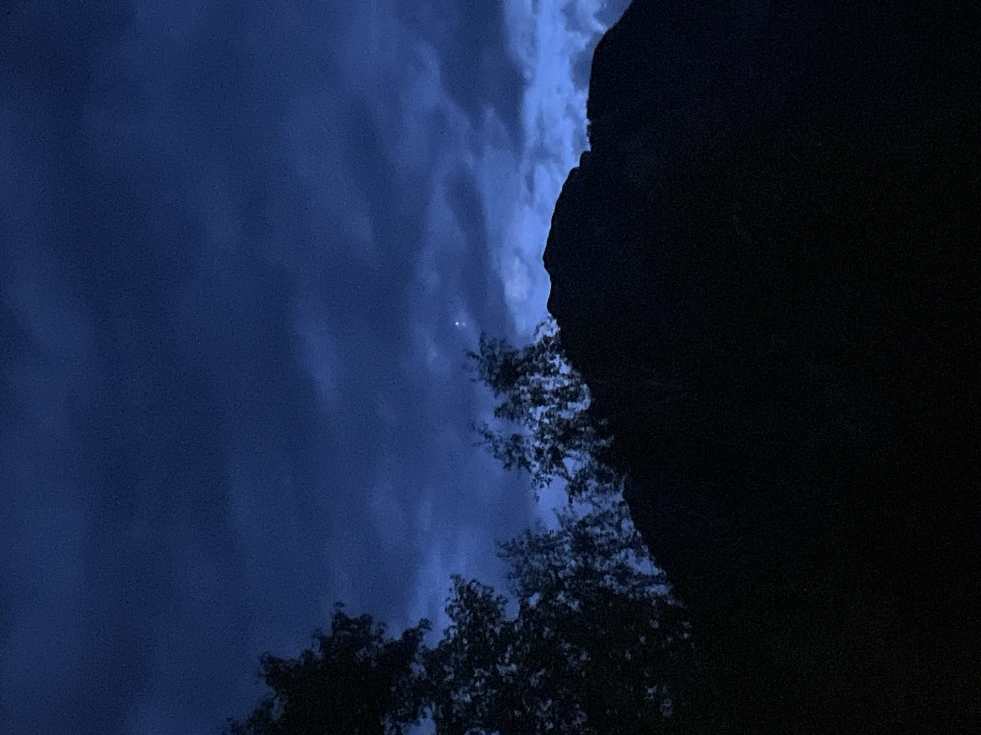 This is a dark, low-light photograph taken in Garmisch-Partenkirchen, Germany, likely during twilight or night. The image features a silhouette of a mountainous landscape against a deep blue, cloudy sky. In the foreground, dark trees with visible branches frame the left and center portions of the image. A prominent, dark silhouette of a mountain ridge occupies the bottom half of the frame. A single, faint star or distant light is visible in the sky, positioned above the mountain ridge and slightly to the left. The overall impression is one of a serene, natural environment under a twilight sky.