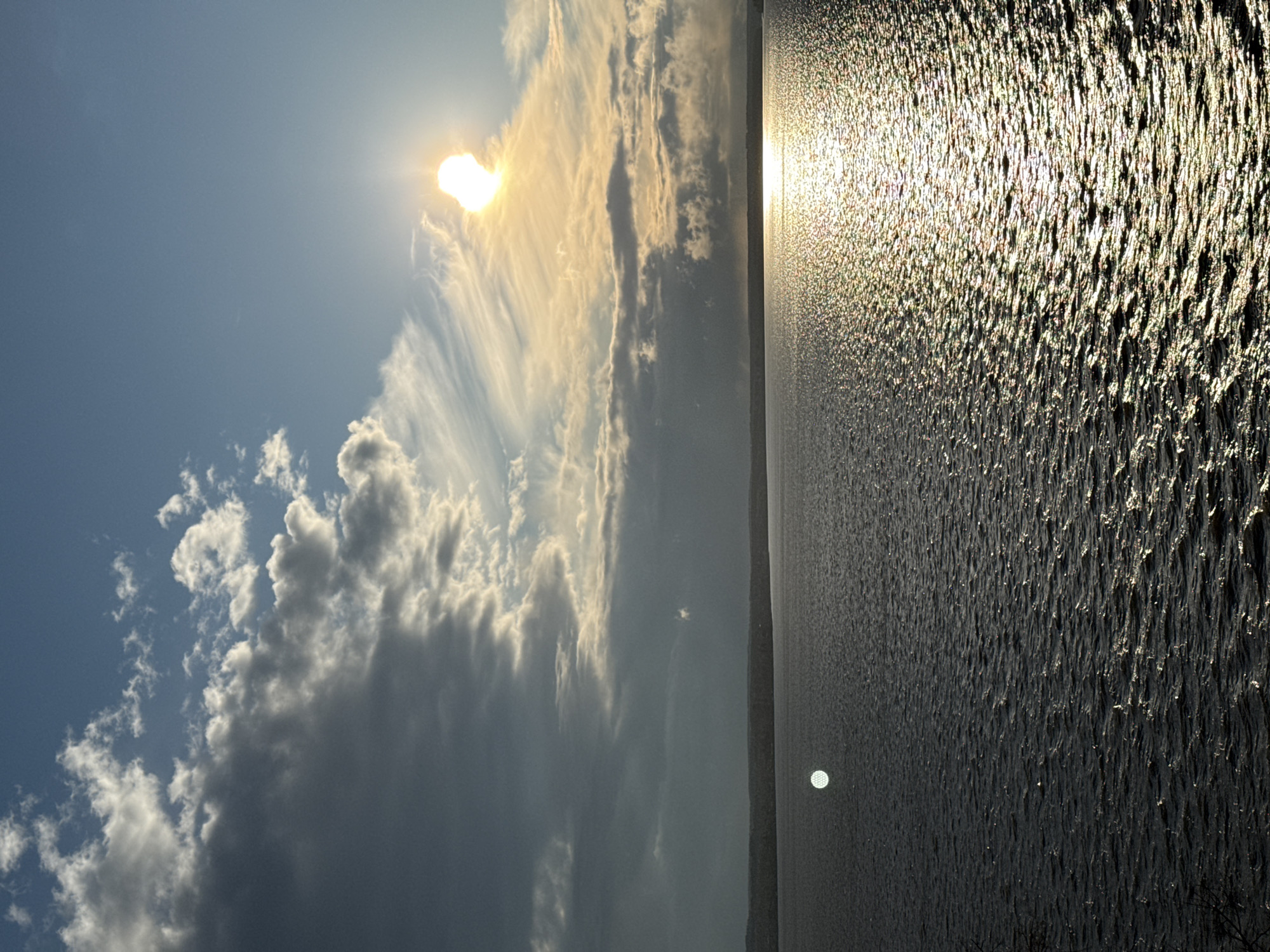 The image shows a landscape with a body of water, likely a lake or reservoir, filling the right half. The water's surface is textured with ripples and reflects sunlight, creating a sparkling effect. On the left side, the sky is filled with clouds, ranging from light and wispy to darker and more substantial cumuliform. The sun is visible behind the clouds, casting a golden glow. The image has a vertical orientation, with the horizon line appearing to be slightly off-center. Given the prompt's specification, this scene is likely in Votkinsk, Russia, presumably at a lake or the Votkinsk Reservoir. There are no people or specific objects visible beyond the natural environment.