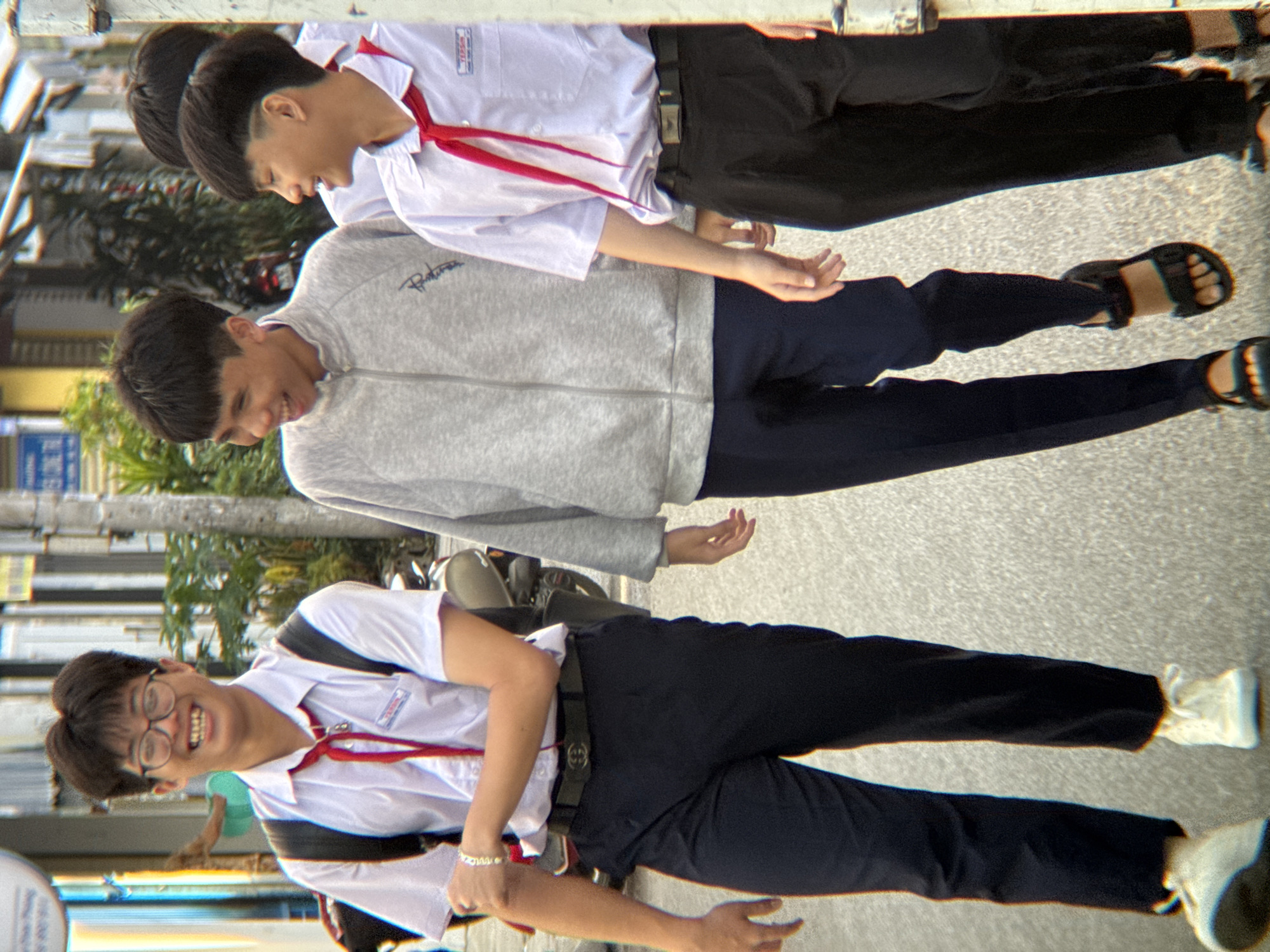 The image shows three young Vietnamese males walking on a paved street, likely in Nha Trang, Vietnam. They appear to be students, all wearing white short-sleeved shirts, dark pants, and red neckerchiefs, suggesting a school uniform. The male in the foreground on the left is smiling and wearing glasses, with a backpack slung over his shoulders. The middle male is wearing a grey zippered jacket over his uniform. The male on the right is also smiling and wearing a red neckerchief. The background shows a street scene with buildings, foliage, and a motorcycle, indicative of an urban environment. The lighting suggests it is daytime.