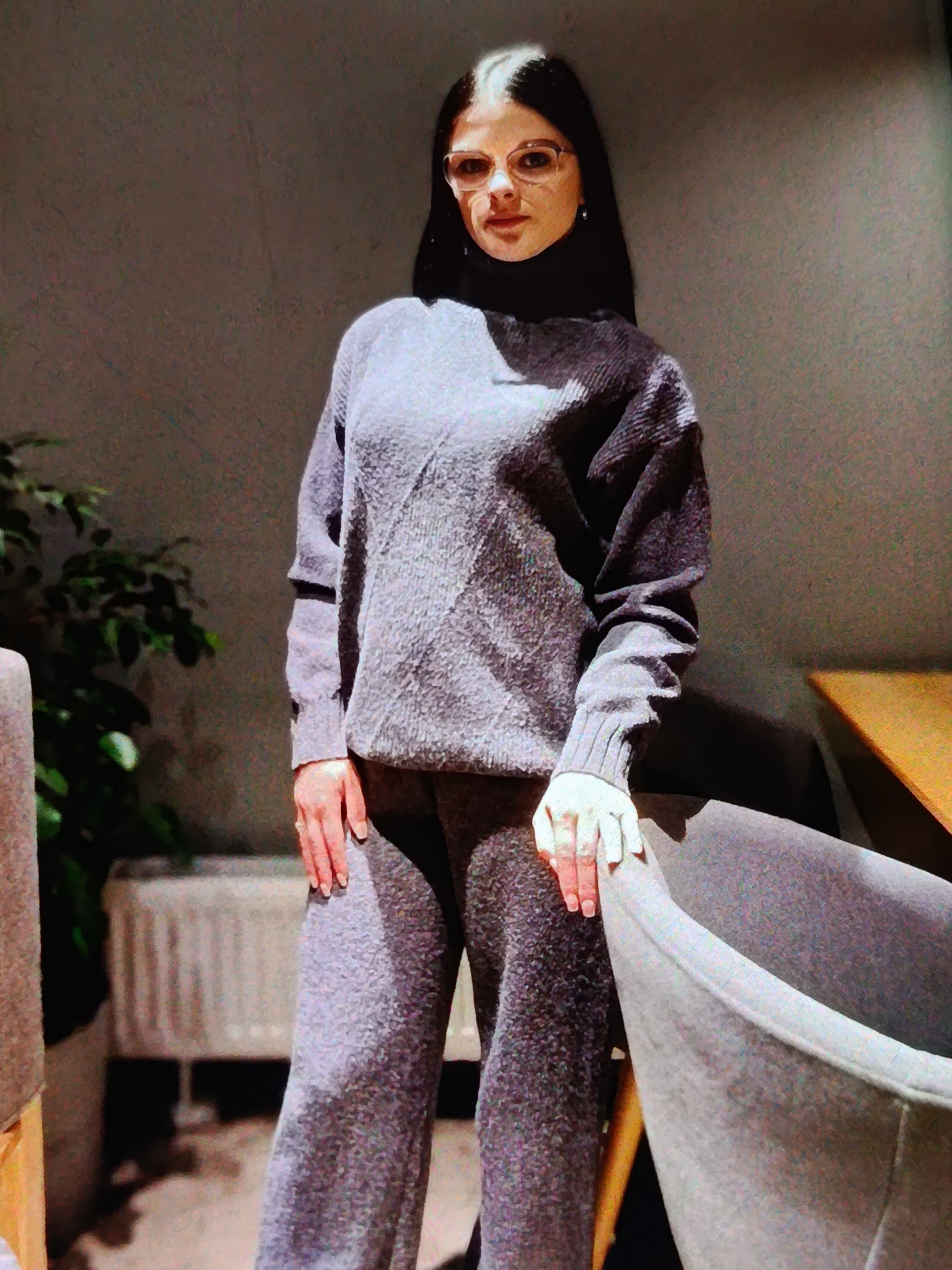 A young woman with dark hair and glasses stands in a dimly lit room, looking directly at the camera. She is wearing a matching grey knit sweater and pants set. The sweater is a turtleneck with a textured knit pattern. Her left hand rests on her leg, and her right hand is visible near her hip, both with manicured nails.
The background is a muted grey wall with a subtle texture. To the left of the woman, a large green plant in a pot is visible. In the foreground, a portion of a grey armchair and a glimpse of a wooden table suggest an indoor setting, possibly a cafe or lounge. A white radiator is visible behind the plant. The lighting is warm and casts shadows, contributing to a cozy, indoor ambiance. The time of day is not explicitly indicated, but the lighting suggests it could be evening or a dimly lit indoor space. No text is visible in the image. The woman's expression is neutral.