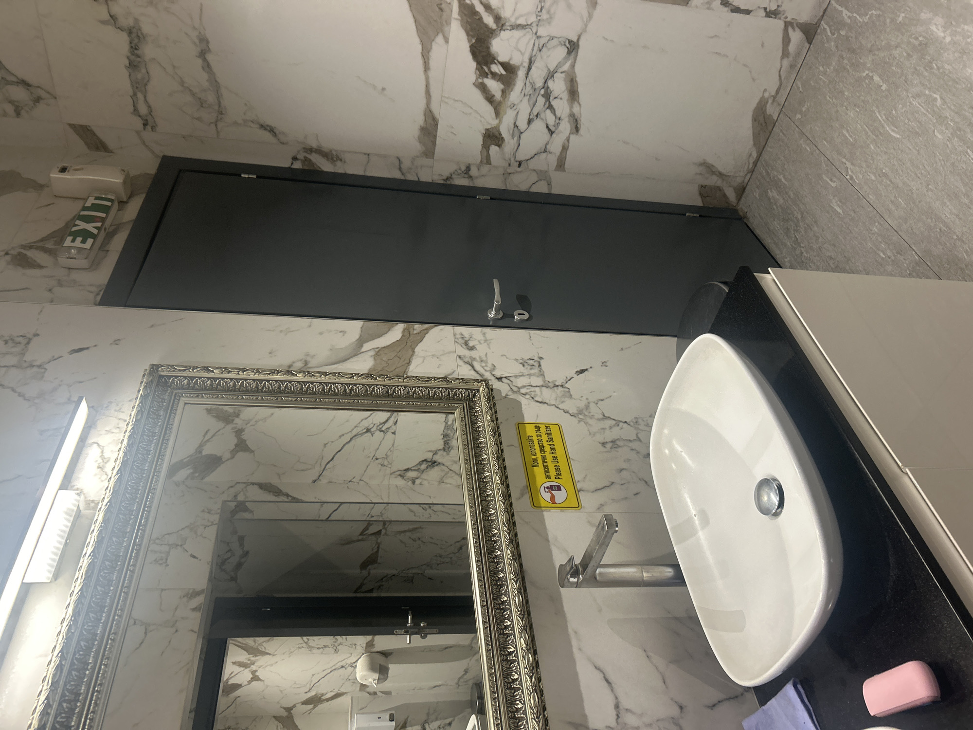 The image shows a modern bathroom with marble-patterned walls and grey tiled floors. A white vessel sink with a chrome faucet is centrally located, placed on a black countertop with a white vanity below. To the left of the sink, a purple cloth and a pink bar of soap are visible on the countertop. A large, ornate mirror with a silver frame hangs above the sink. To the right of the mirror, a dark grey door is visible, with a silver handle and lock. Above the door, an illuminated "EXIT" sign is mounted on the wall. A yellow sticker with text in Bulgarian and English, reading "Molia, izpolzvaite antiseptichno sredstvo za raka. Please Use Hand Sanitizer," is affixed to the mirror. The overall impression is of a clean and well-maintained restroom.