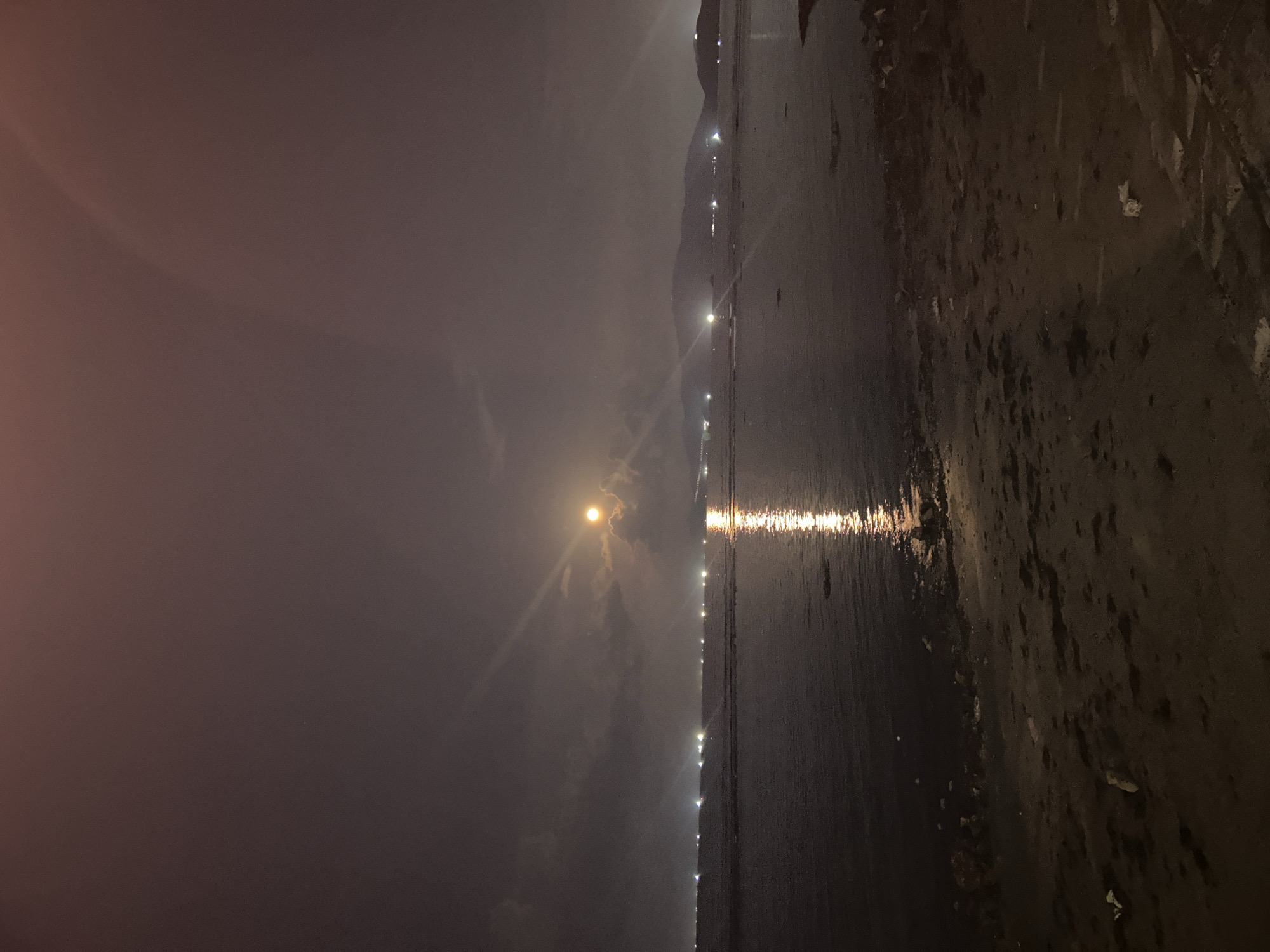 The image depicts a nighttime view of a beach in or near Bac Nha Trang Ward, Vietnam. A bright moon is visible in the sky, partially obscured by clouds. A line of lights, likely from buildings or street lamps, stretches along the coast. The moon's reflection creates a bright streak on the water's surface. The beach itself appears dark and wet.
