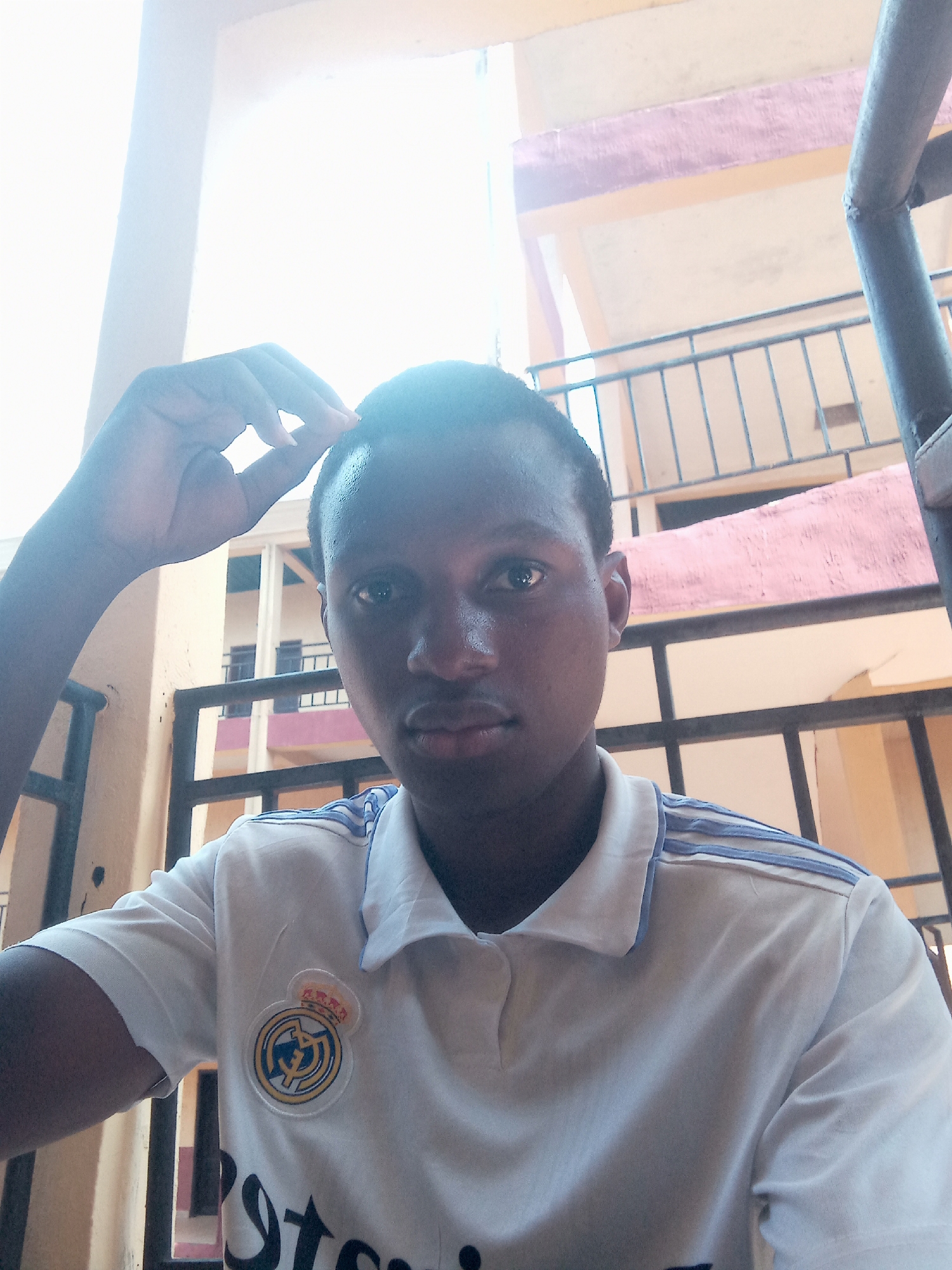 A young Black male is captured in a close-up, portrait-oriented photograph, looking directly at the camera with a neutral expression. He appears to be in his late teens or early twenties and has short, dark hair. His right hand is casually raised, with fingers lightly touching his forehead.
He is wearing a white short-sleeved polo shirt that features light blue stripes on the shoulders and a circular Real Madrid crest prominently displayed on the left side of his chest. The lower part of a dark logo, consistent with the "Emirates" sponsor often found on Real Madrid jerseys, is partially visible towards the bottom of his shirt.
The setting appears to be outdoors, possibly on a balcony or an open-air corridor of a building. Metal railings are visible behind him, and the background shows parts of a building structure with walls painted in beige and a lighter, somewhat faded pinkish-purple hue. Bright natural light illuminates the scene from above, causing some overexposure in the upper portion of the background, suggesting clear, sunny daytime conditions. The overall image presents a casual portrait in what appears to be an urban or residential environment in Nkwelle, Nigeria.