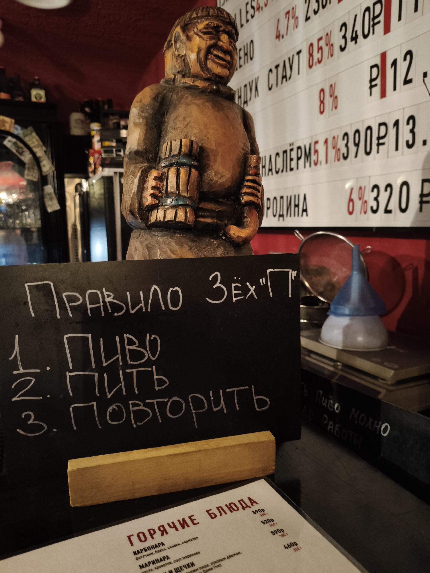 A close-up shot depicts a bar or pub setting, likely in Chaikovsky, Russia, given the Cyrillic script on signs and menus. A wooden carving of a jovial-looking monk-like figure holding a beer stein dominates the foreground. In front of the figure, a small chalkboard displays the humorous "rule" in Russian: "ПРАВИЛО ЗЁХ 'П'," followed by "1. ПИВО" (Beer), "2. ПИТЬ" (Drink), and "3. ПОВТОРИТЬ" (Repeat). A wooden stand supports the chalkboard, and a menu titled "ГОРЯЧИЕ БЛЮДА" (Hot Dishes) is visible below, listing dishes like "КАРБОНАРА" (Carbonara) and "МАРИНАРА" (Marinara) with prices in rubles.
Behind the wooden figure, a white board is visible displaying the price and percent alcohol by volume (ABV) of various beer types: "LES 4.7% 23," "EHO НДУК СТАУТ 8.5% 340P," "8% P12," "НАСЛЁРМ 5.1% 390Р13," "РОДИНА 6%320 P".
Further in the background, bottles and other bar paraphernalia can be seen. There is a strainer and a blue and white funnel on top of a book. A chalkboard on the right has illegible writing, but mentions "Пиво МОЖНО ДО РАБОТЫ" (Beer allowed before work.) The lighting is dim.