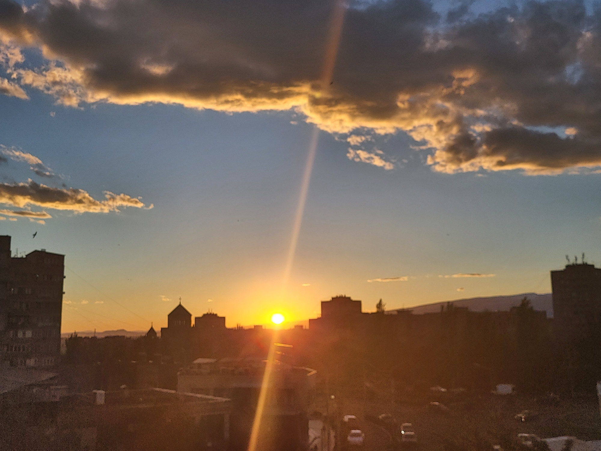The image captures a sunset view over Yerevan, Armenia. The sky is a gradient of blue and orange, with clouds illuminated by the setting sun. Silhouetted buildings, including what appears to be a church with a dome, define the city skyline. A bright sun sits on the horizon, casting a strong ray of light downwards. The foreground shows rooftops and streets with a few cars visible. The overall scene is a cityscape at dusk.