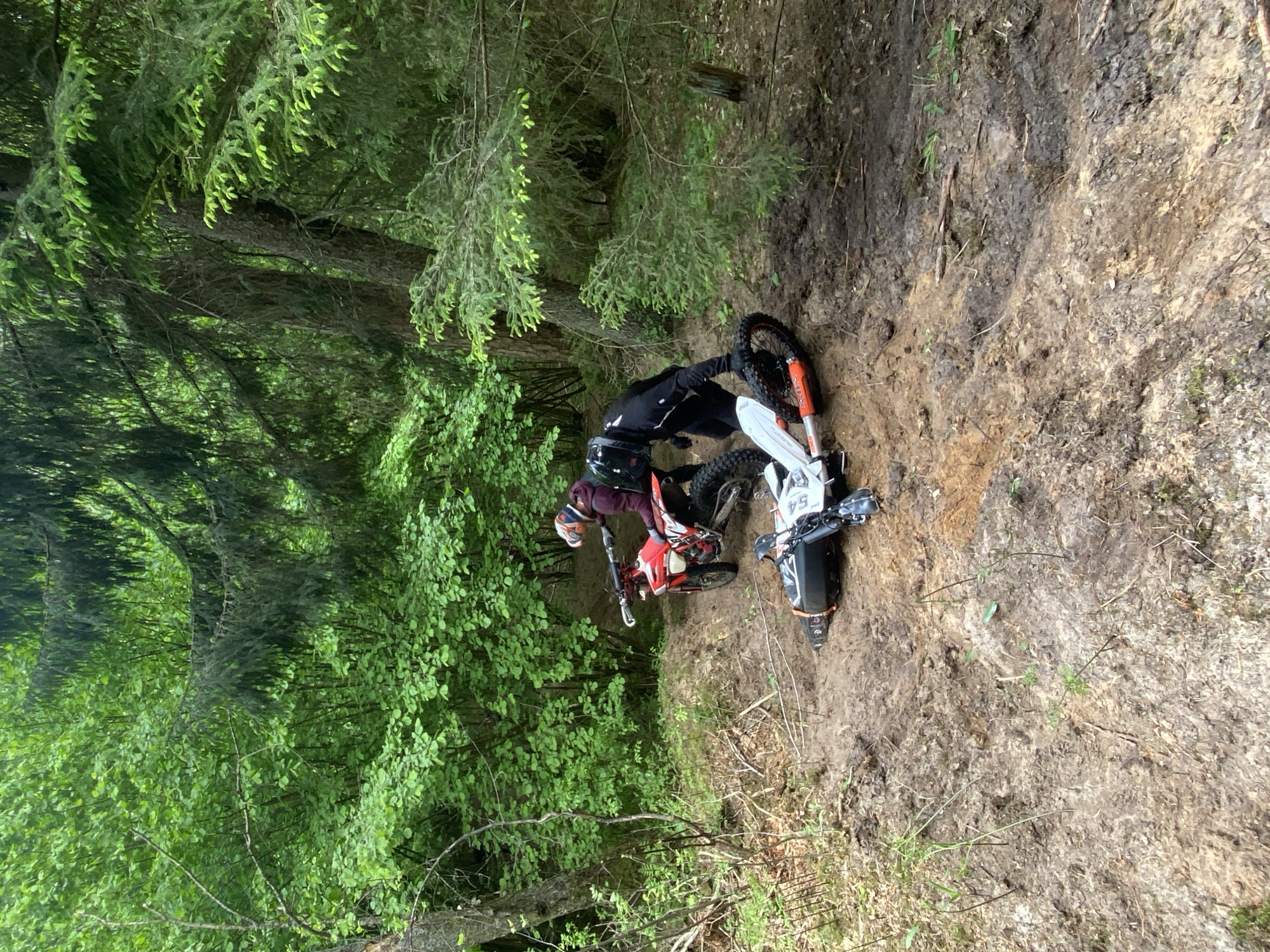 The image depicts a person with a helmet, wearing black pants and a dark-colored shirt, standing near two motorcycles in a forested area. One motorcycle is red, and the other appears to be white with black accents. The location appears to be a hillside or embankment with a dirt surface. The environment is heavily wooded, and the picture was taken in Парэцкі сельскі Савет, Belarus.