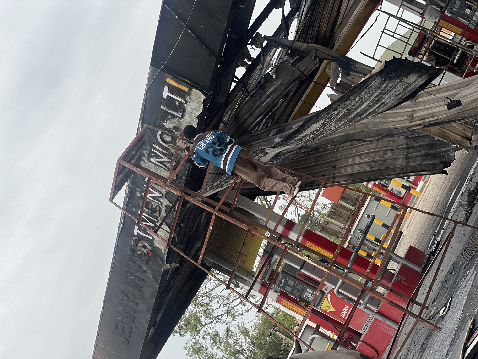 A man wearing a blue jersey and brown pants stands on a metal ladder, positioned in front of a damaged structure with the name "JENNY INVESTMENT NIGERIA LTD" partially visible. The scene depicts the aftermath of a fire, with charred and twisted metal roofing, likely at a petrol station in Gura Top, Nigeria. Red petrol pumps are visible in the background. The sky is overcast.