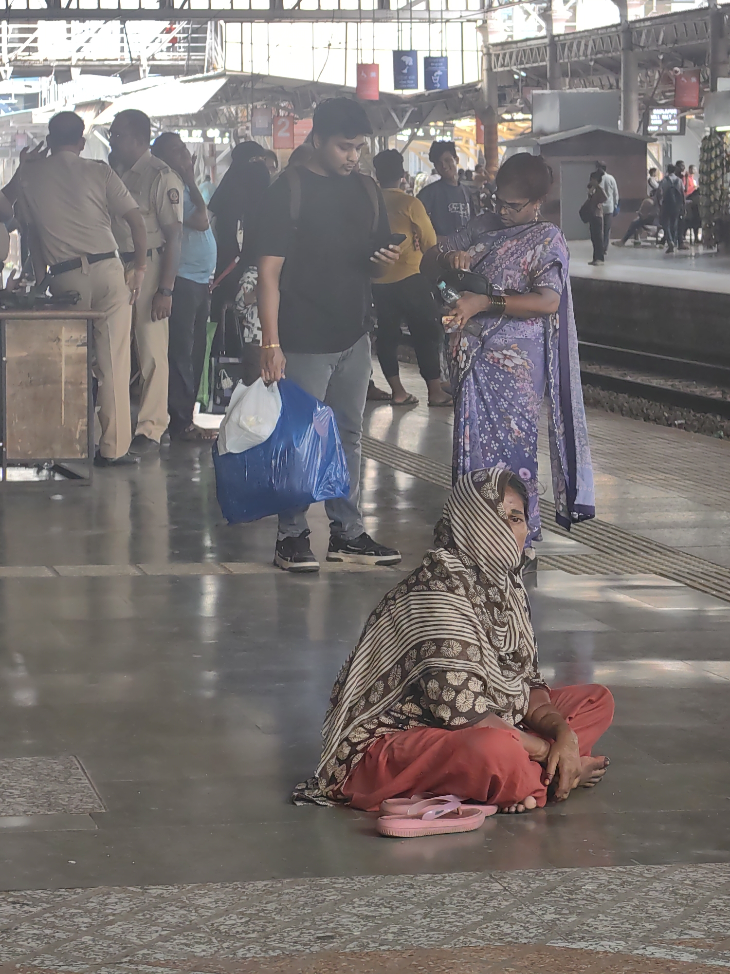 The image depicts a busy scene on a train platform, likely in Mumbai, India, given the context of the architecture and people's attire. In the foreground, a woman is seated on the ground, wrapped in a patterned shawl that covers her head and shoulders, with her legs crossed and wearing orange trousers. A pair of pink flip-flops are placed to her left.
Behind her, a woman in a purple floral sari stands, holding a black purse and looking down as if searching for something. Next to her, a man in a black t-shirt and grey pants is also looking down, possibly at his phone. Further to the left, two uniformed police officers stand near a wooden counter. One officer appears to be speaking to someone off-camera, while the other faces away from the viewer. A blue plastic bag and a white bag are held by another individual whose legs are visible. In the background, various other individuals are present, suggesting a typical day at a bustling train station with people waiting for trains, passing through, or engaged in conversations. The platform is paved with concrete, and tracks are visible to the right. The overall atmosphere is one of movement and activity within the urban transit environment.