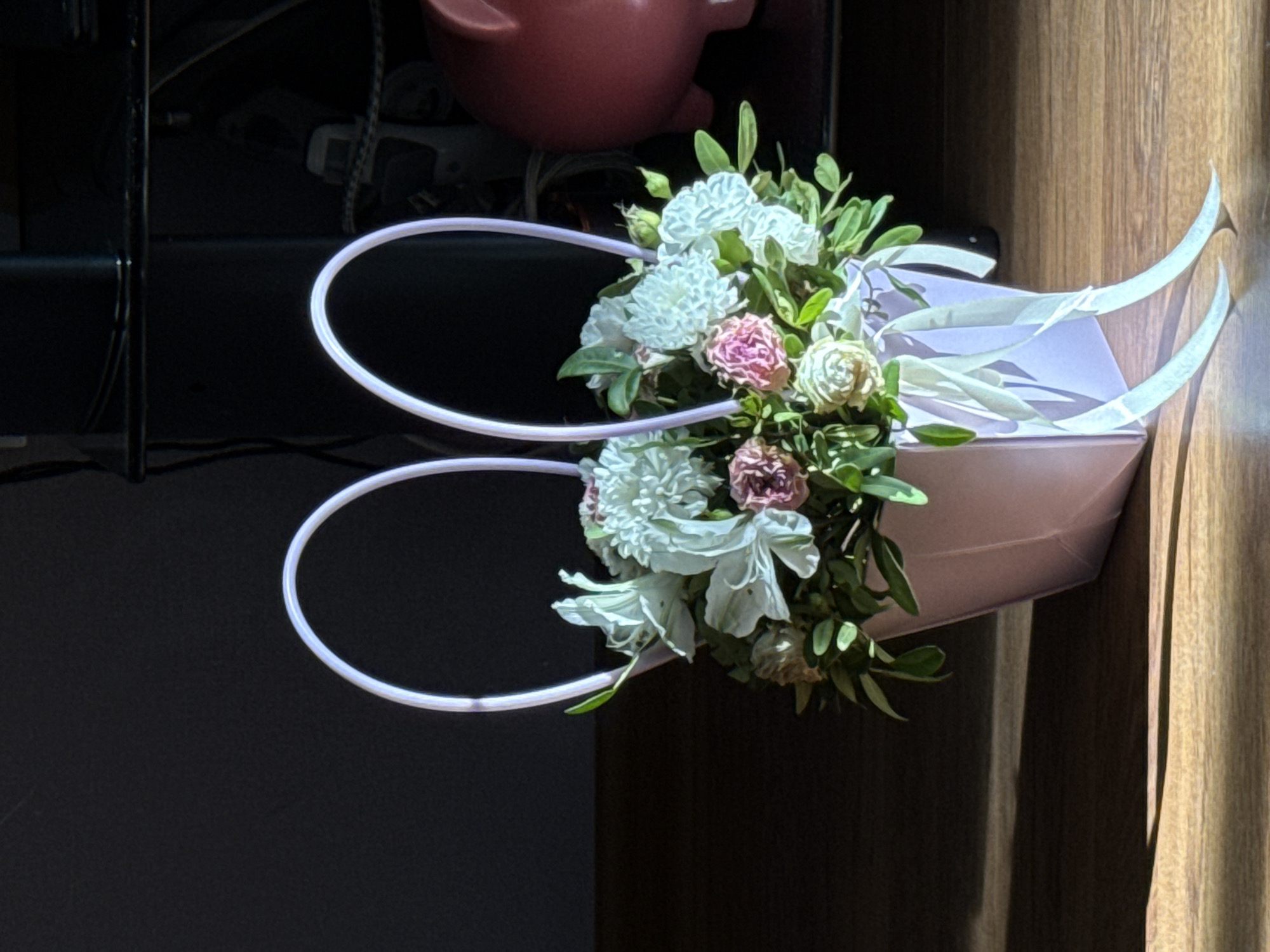 The image shows a pale purple, two-handled basket filled with a floral arrangement. The arrangement consists of white, pink, and off-white flowers, interspersed with green foliage. The basket appears to be positioned indoors, near a wooden-paneled wall on the right side of the frame, with a dark background on the left. The lighting is bright on the right side, with a shadow casting across the lower left. There is no indication of the location being in Chaikovsky, Russia, or any people present.