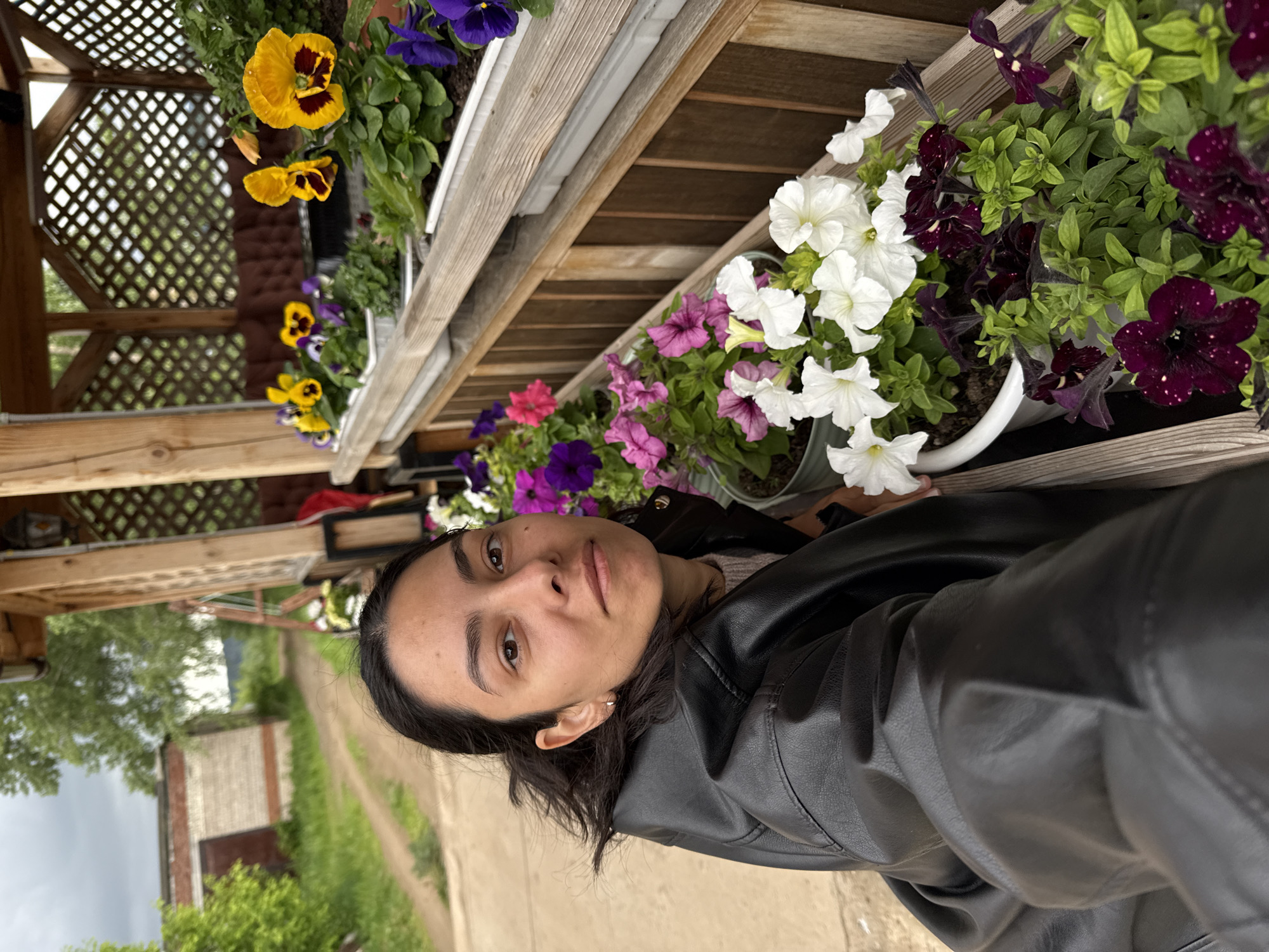 A young adult female is depicted in a close-up, taking a selfie or being photographed, positioned alongside a rustic wooden gazebo in Chaikovsky, Russia. She has dark hair, dark eyes, and wears a black leather jacket over a light-colored knitted top, looking directly at the camera with a neutral expression. A small, silver hoop earring is visible in her left ear. The wooden gazebo features a lattice-style fence and supports multiple white planters filled with various blooming flowers, including yellow and purple pansies, and pink, bright purple, white, and dark maroon/black petunias. In the background, a dirt road or path extends into the distance on the left side of the frame, bordered by green foliage. A low, light-colored brick building with a darker roof and door is visible to the left of the path, suggesting a garage or shed. The sky is overcast.