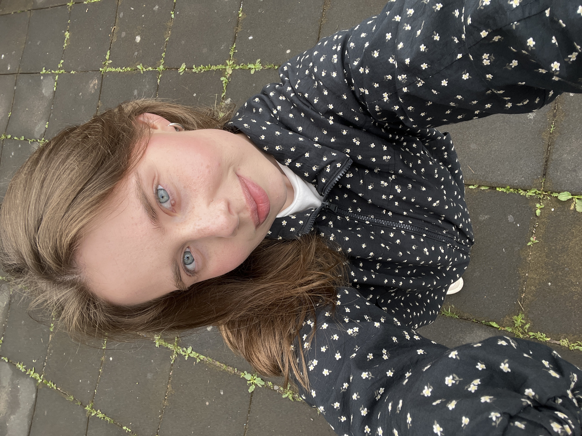 A young woman is captured in a selfie taken from an overhead perspective, looking directly up at the camera. She has long light brown hair, fair skin, and piercing light blue eyes. Freckles are visible across her nose and upper cheeks, and she has a soft, friendly smile. She is wearing a black jacket or top patterned with numerous small white and yellow flowers, featuring a prominent black zipper down the front. A simple white shirt is visible underneath. A small silver hoop earring adorns her right ear.
The setting is outdoors, with the background consisting of dark grey square or rectangular paving stones. Green weeds and small plants are growing in the gaps between the pavers. Some sections of the pavement appear slightly damp, suggesting recent rain or humid conditions. The overall lighting indicates it is daytime, likely overcast. This scene is set in Chaikovsky, Russia. There is no visible text in the image.