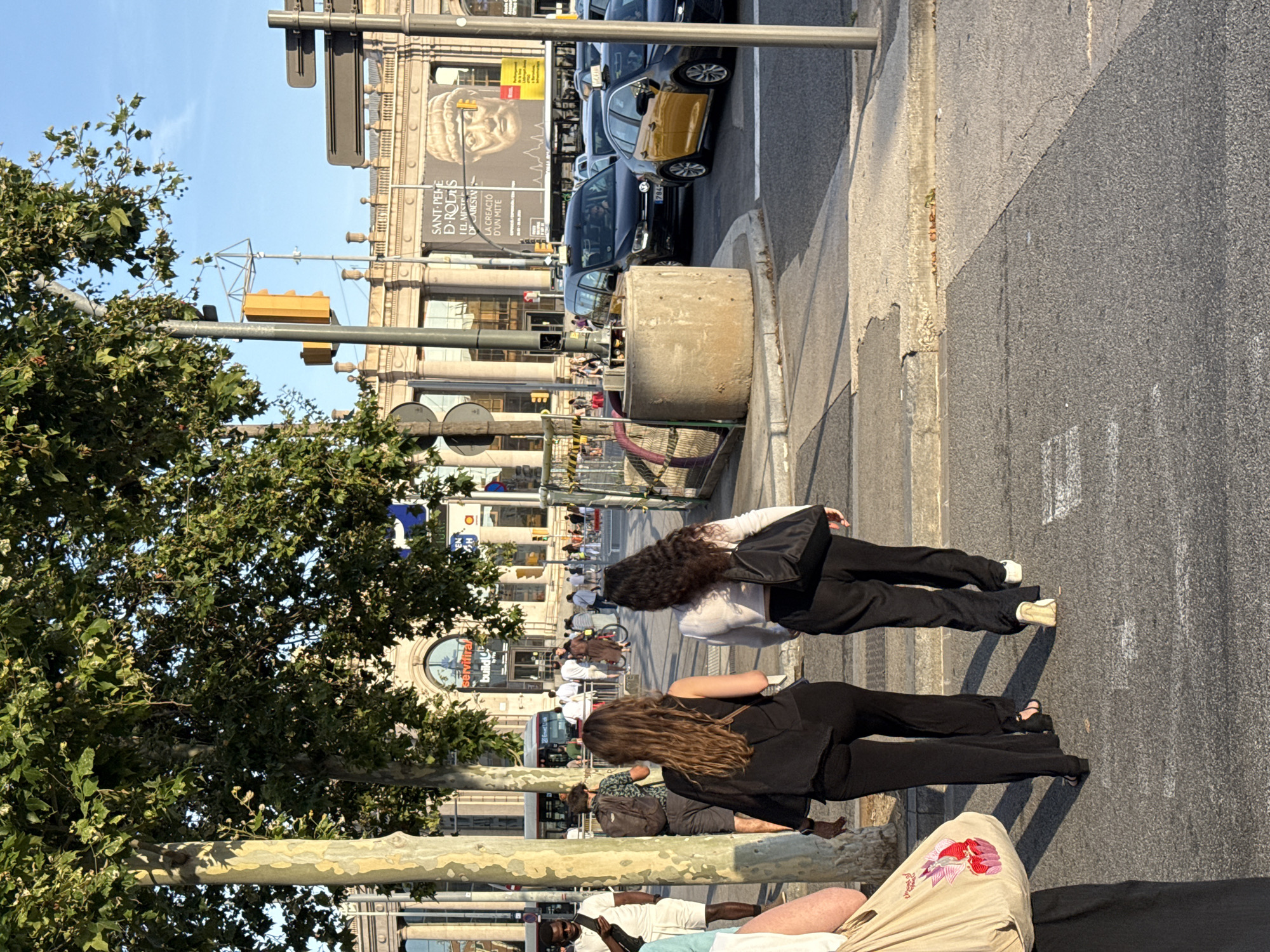 Two women are walking away from the camera on a pedestrian crossing. The woman on the left has long brown hair and is wearing a black top and black pants. The woman on the right has dark curly hair and is wearing a white top and black pants. Both women are carrying black bags. In the background, there are several cars, including two yellow taxis, and a bus. A large building with columns is visible behind the cars. The building has a banner with a large image of a Roman bust and text in Catalan. The scene appears to be set in the city of Barcelona, Spain, likely near a transportation hub or a historical site, given the architecture and the presence of the banner. The sky is clear and blue, suggesting it is a sunny day.