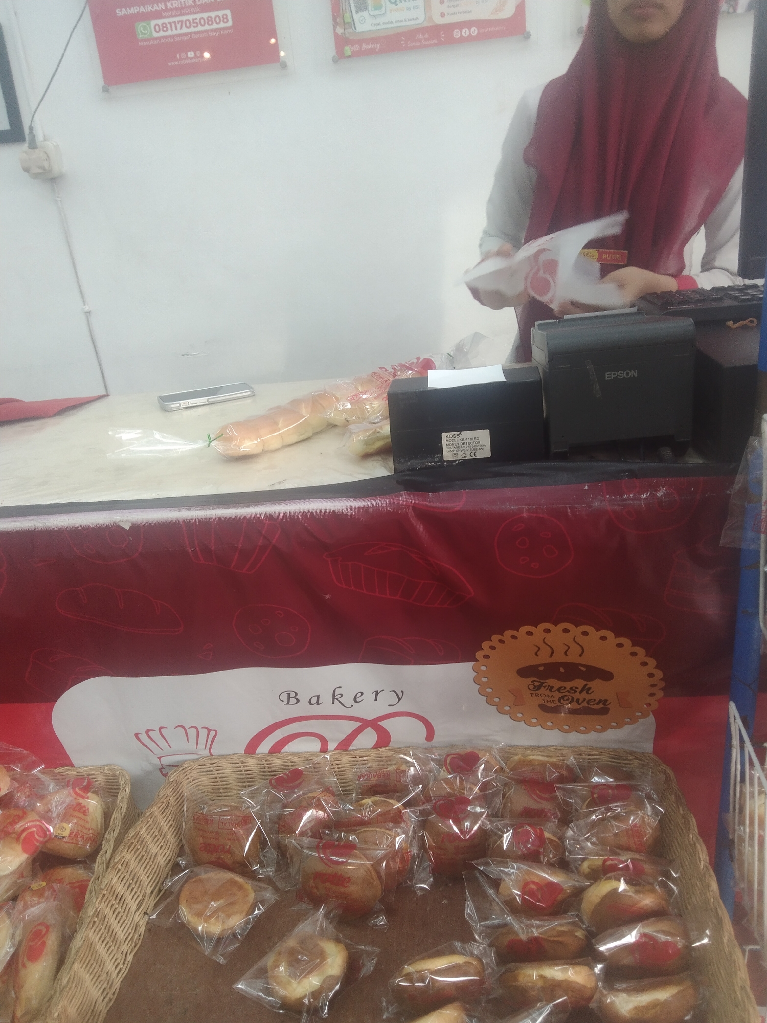 A person wearing a maroon hijab is standing behind a counter. In front of the person, there is a cash register with an Epson printer attached, and another electronic device with a label that reads "KOSS". A loaf of bread is visible on the counter, partially wrapped in plastic. In the foreground, there are two wicker baskets filled with individually packaged baked goods. A red banner with white illustrations of baked goods and the word "Bakery" is displayed behind the baskets. A sign with "Fresh From the Oven" is also visible on the banner. The environment appears to be a bakery in Siak Sri Indrapura, Indonesia.