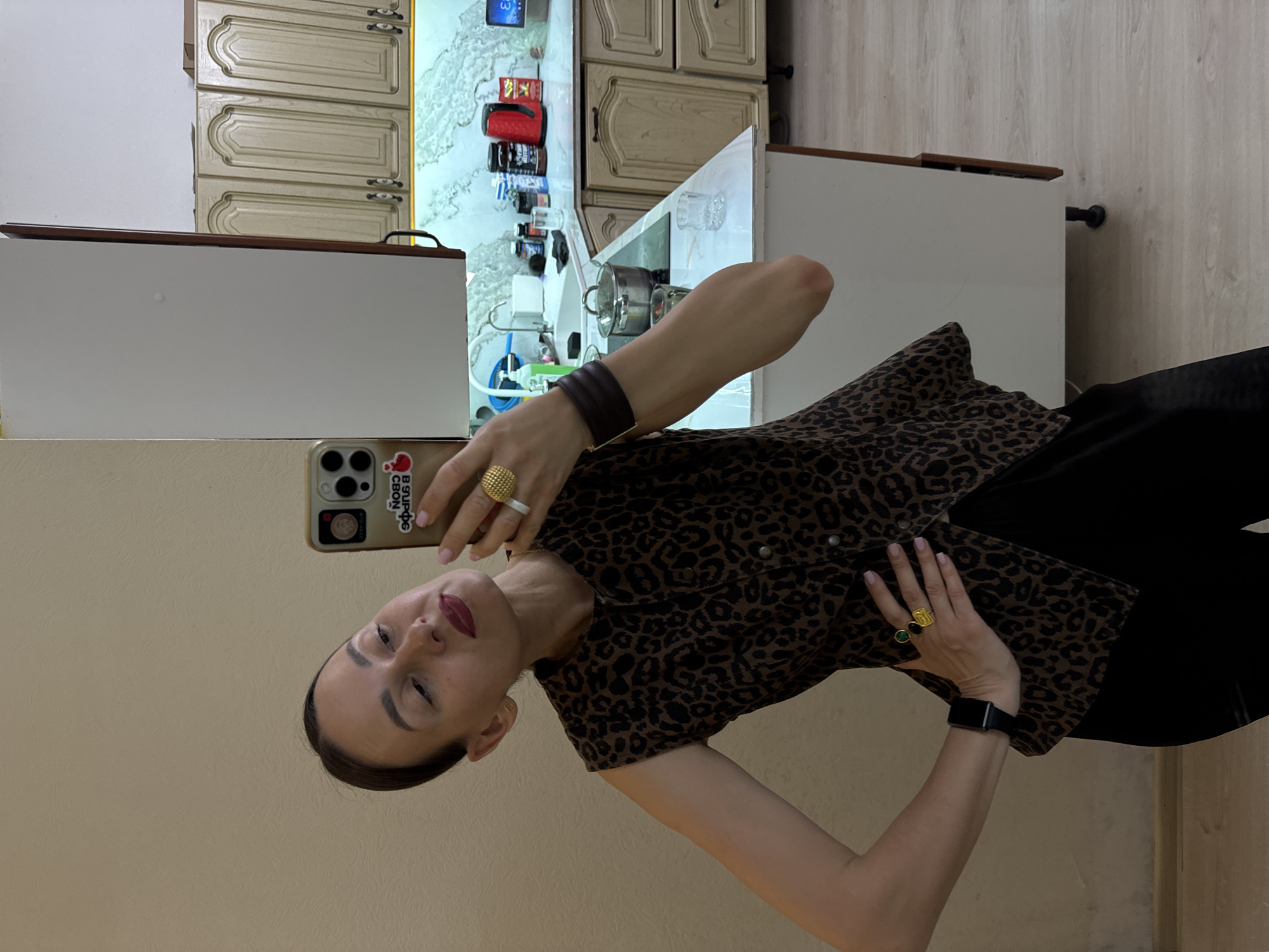 A woman is taking a mirror selfie in what appears to be a kitchen. She is wearing a sleeveless leopard-print top and black pants, with several rings on her fingers and a watch on her left wrist. The kitchen features light-colored cabinets, a marble countertop, and a sink. The overall environment suggests an indoor residential setting. There is no indication that this scene is in Sochi, Russia.