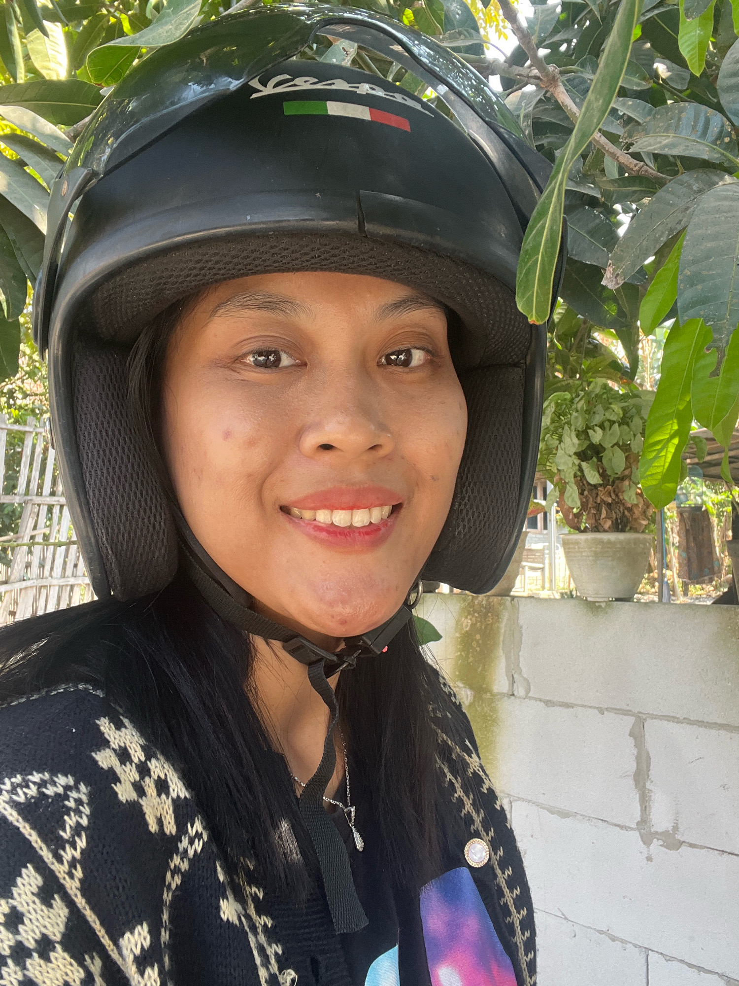 A woman wearing a black helmet and a black sweater with a geometric pattern poses for a selfie outdoors. She has dark hair and a light complexion, with a smile revealing white teeth. The background shows lush green foliage, a white brick wall, and some potted plants. A bamboo fence is visible to the left of the woman. The image appears to be taken during the daytime, with natural sunlight illuminating the scene. The woman's attire and the surrounding environment suggest a casual, possibly rural or suburban setting in Kecamatan Pati, Indonesia.