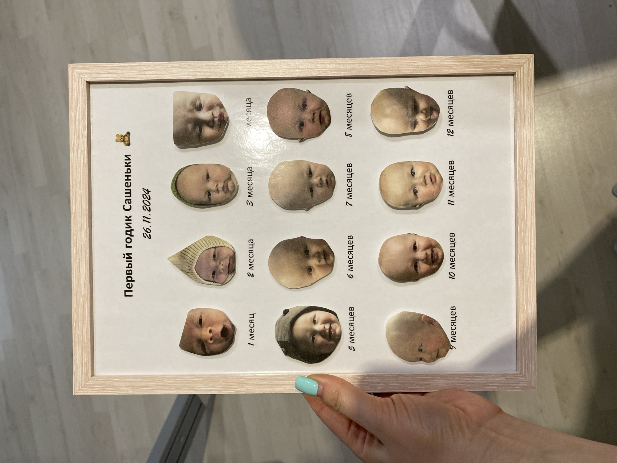 A light-skinned hand with a light blue-green painted thumbnail holds a vertical, light wood-framed picture. The picture displays a white background with Cyrillic text at the top reading "Первый годик Сашеньки" (Sashenka's First Year) and the date "26.11.2024." A small teddy bear emoji is positioned next to the text. Below the text, twelve circular or oval-shaped headshots of a baby are arranged in four rows of three, depicting the baby's growth. Each photo is labeled in Cyrillic with a corresponding month, from "1 месяц" (1 month) to "12 месяцев" (12 months). The background is an out-of-focus indoor environment with a light-colored floor, possibly laminate or tile, and a light wall or surface in the upper left. The presence of Cyrillic text indicates the scene is located in a Russian-speaking region, consistent with the city of Izhevsk, Russia.