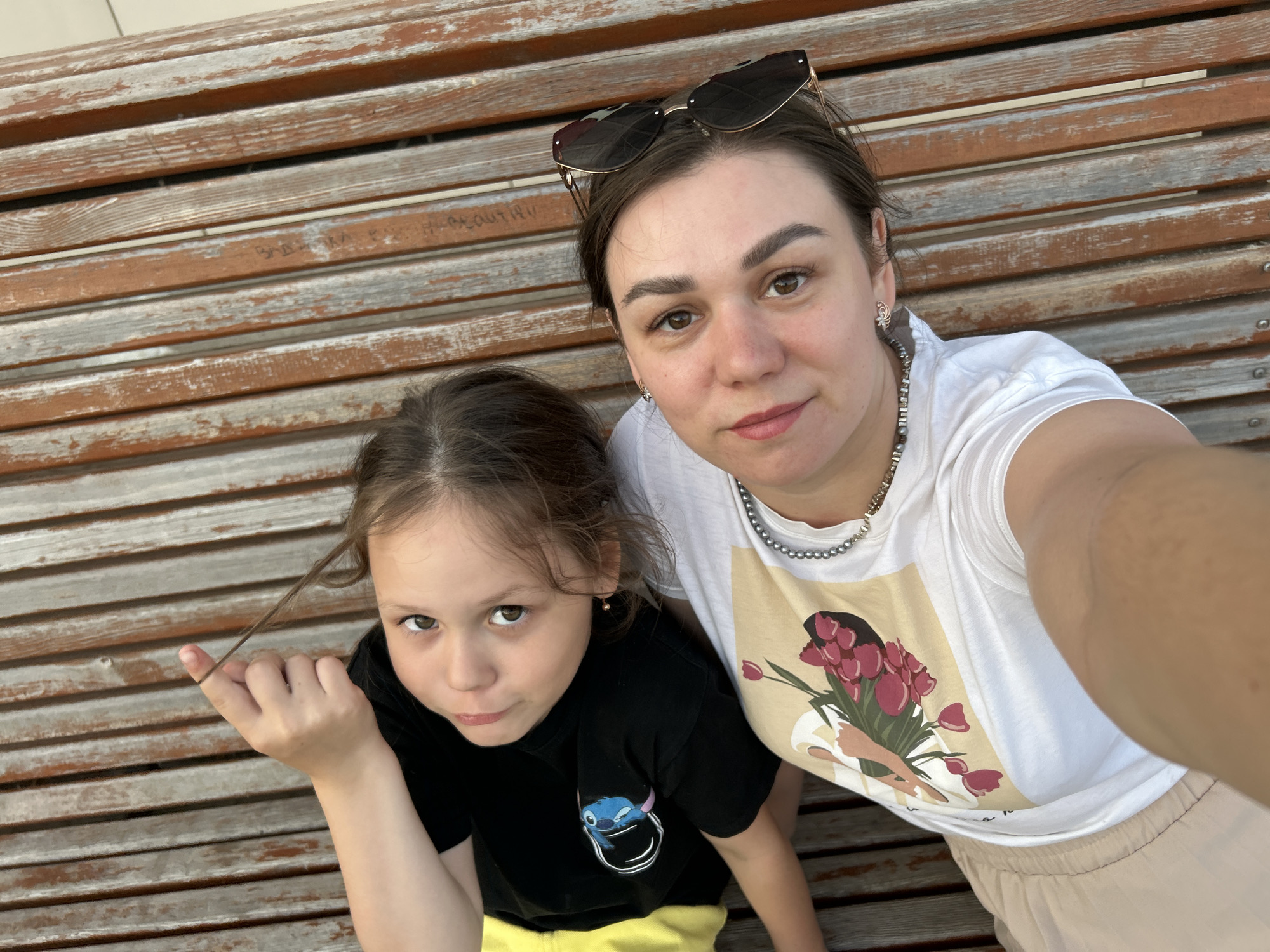 A woman and a child are captured in a selfie taken on a wooden bench. The woman, sporting sunglasses atop her head, a white T-shirt with a floral design, and a pearl necklace, is positioned slightly behind the child. The child wears a black T-shirt and yellow shorts, holding a strand of hair. The background reveals a weathered wooden bench. The location is Chaikovsky, Russia.