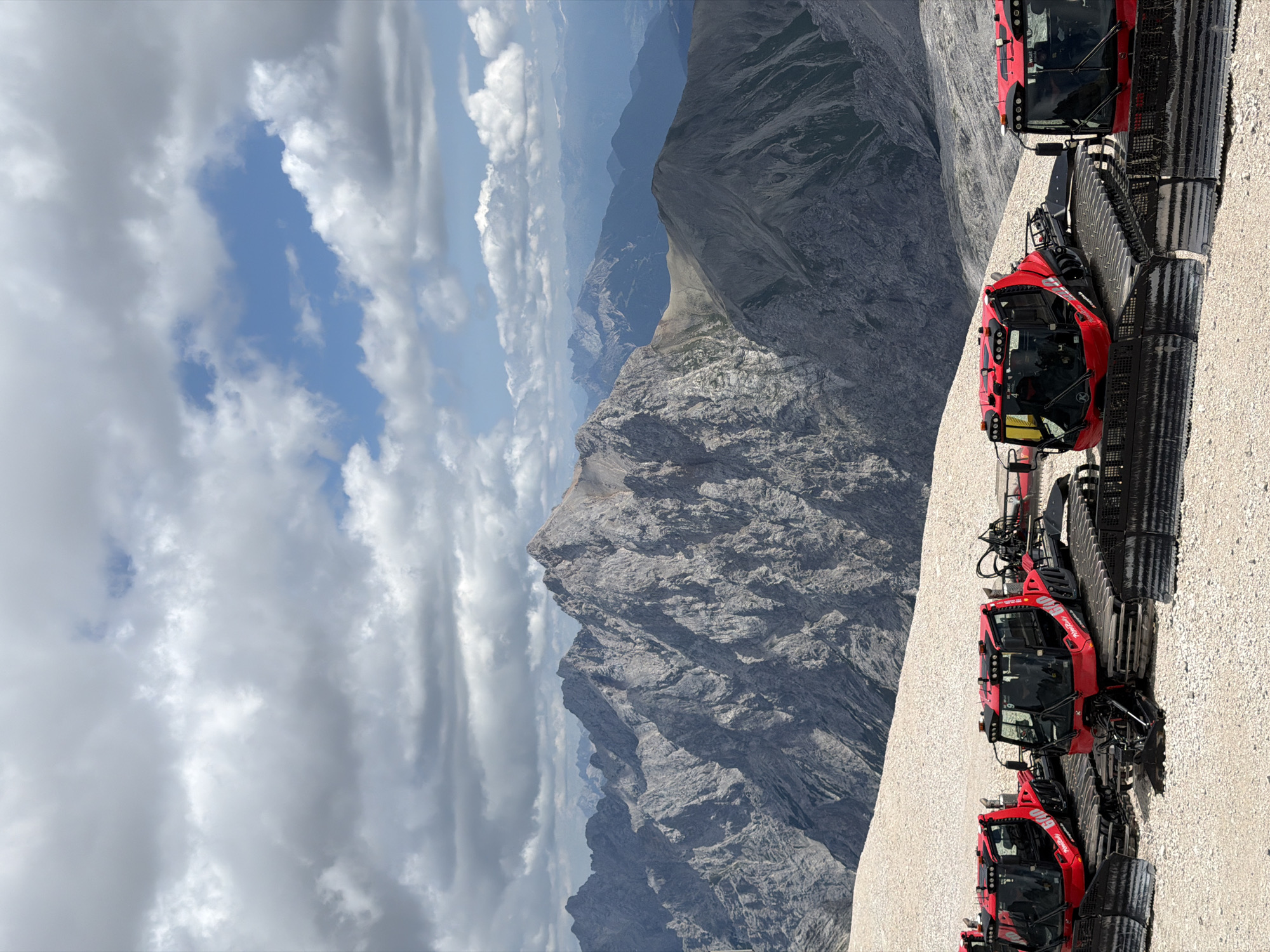 The image depicts a panoramic view of the Bavarian Alps, likely from the Zugspitze, Germany's highest peak, near Garmisch-Partenkirchen. In the foreground, a line of four red snowcats is parked on a gravelly, snow-dusted slope. These vehicles are equipped with large tracks and front plows, suggesting they are used for grooming ski slopes or maintaining winter trails.
The middle ground is dominated by rugged, gray, rocky mountain ranges with sharp peaks and deep valleys. The texture of the rock formations is detailed, showing signs of erosion and weathering. In the background, a series of progressively lighter blue mountain ranges recede into the distance, indicating a vast and expansive mountainous landscape.
The sky above is a mix of bright blue patches and scattered, fluffy white and gray clouds. The lighting suggests it is daytime, with sunlight intermittently breaking through the clouds, casting shadows on the mountains. There are no people visible in the image. The overall impression is one of a remote and majestic alpine environment, prepared for winter activities.