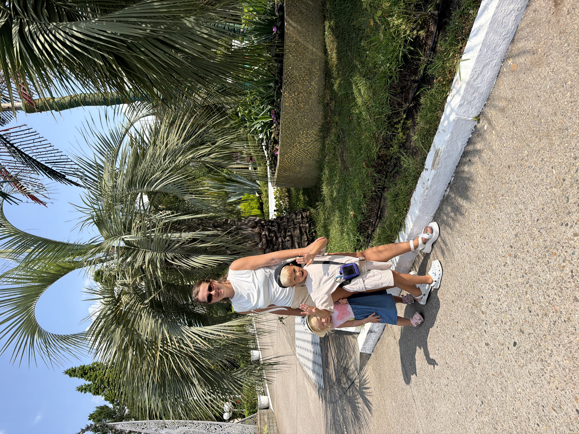 A woman and two young children are standing on a paved path in front of large palm trees. The woman is wearing sunglasses, a white sleeveless top, and light-colored shorts. The boy, on her right, has a black baseball cap on backwards and is wearing a light-colored t-shirt and shorts. He has a small blue bag hanging from his shoulder. The girl, on the woman's left, is wearing a pink shirt with a heart and a blue skirt. She has a straw hat on. The children are both waving. The path is bordered by a white-painted curb and a grassy area with plants and flowers in a decorative, tiled planter. In the background, there are more palm trees and some evergreen trees. The sky is blue and clear. The scene appears to be in a park or botanical garden. The setting is consistent with the arboretum in Sochi, Russia.