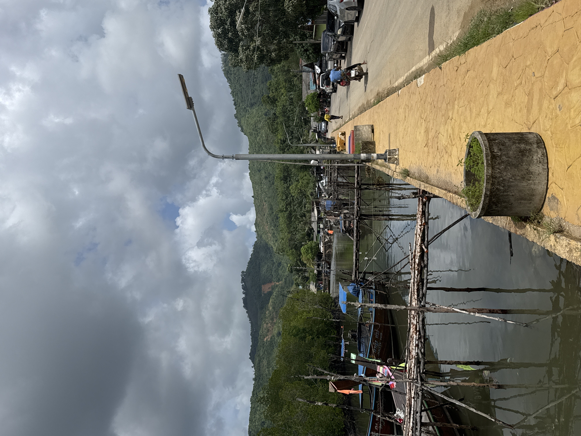 In Ban Hin Rom, Thailand, a cloudy day casts shadows over a waterfront scene. A narrow waterway runs alongside a road, with wooden piers and traditional longtail boats moored on the left. The water reflects the overcast sky and the surrounding mangrove trees. On the right, a paved yellow walkway runs parallel to the road, bordered by sparse grass. A man on a motorcycle rides away from the camera on the road, and parked cars are visible further down. In the background, lush green hills rise, with a noticeable landslide scar on one slope. A street lamp stands prominently on the walkway.