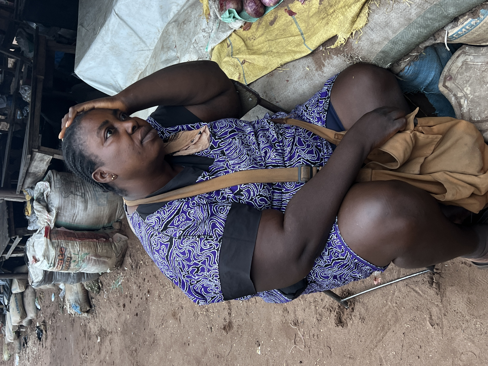 A Black woman is crouched down in an outdoor market in Nkpor, Nigeria. She is wearing a purple and white patterned dress with black trim. A tan strap from a bag is visible across her chest. Her right hand is on her forehead, and she is looking to the side with a pensive expression. The ground around her is made of dirt and scattered debris. Behind her, there are large sacks of what appear to be cement or grain, and a wooden structure that suggests a stall. To her right, a yellow tarp is laid out, with some red onions visible on it. The overall scene depicts a person in a bustling but seemingly low-income market environment.