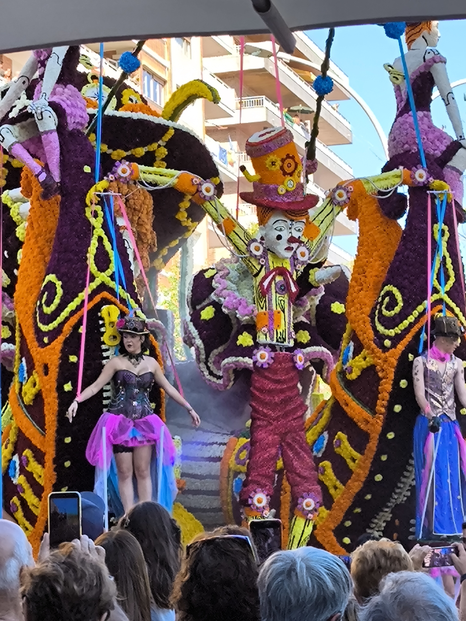 The image captures a vibrant outdoor parade float decorated with abundant colorful flowers, likely part of a festival in Laredo, Spain. The float features a central, larger-than-life figure dressed in a clown-like costume with a tall, ornate hat and brightly colored pants. To the left and right of this central figure are two smaller figures, one appearing to be a performer in a corset and a flowing pink and blue skirt, and another on the far right, dressed in a patterned vest and a blue skirt. These figures are integrated into the elaborate floral decorations that form butterfly-like wings and vertical structures. A crowd of spectators is visible in the foreground, holding up phones to capture the spectacle, indicating a public event. The background reveals buildings with balconies, suggesting an urban setting. The lighting suggests it is daytime.