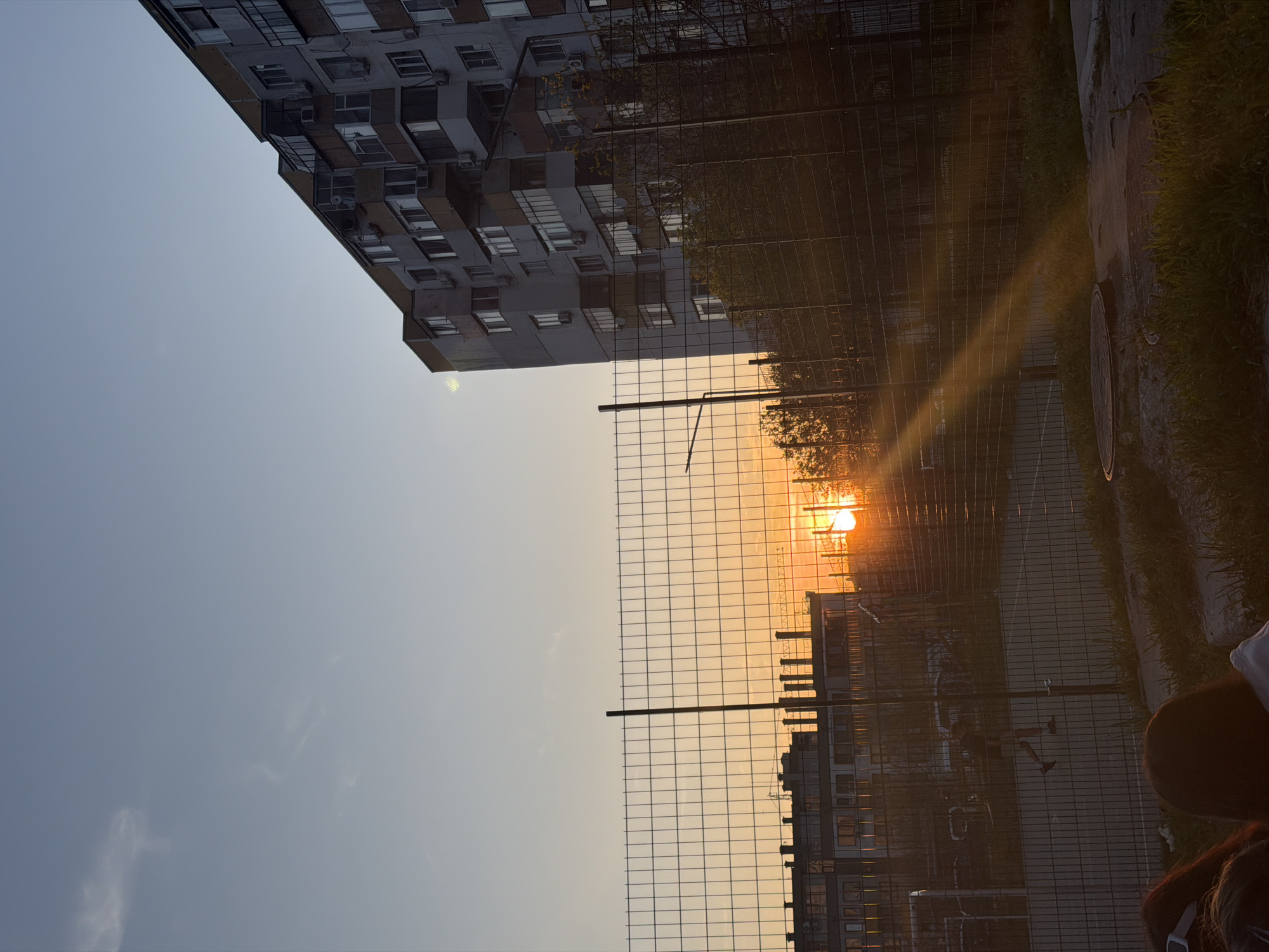 The photograph appears to be taken outdoors, near a residential area in Varna, Bulgaria. A tall apartment building is visible in the upper left corner of the frame. A metal chain-link fence runs across the majority of the image. The sun is setting, creating a bright orange glow in the distance, partially obscured by the fence. The light from the sunset has cast a ray across the frame. Part of a manhole cover can be seen next to grass in the upper right. In the lower right, a head is visible, indicating the presence of people, likely the photographer. The general atmosphere is one of evening, possibly late summer or early fall. The location is likely a park or a space next to the residential building in a city where construction is apparent.