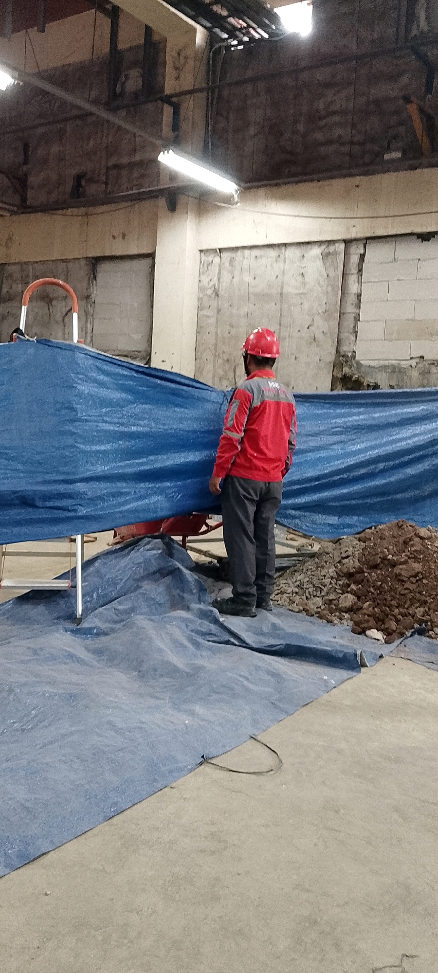 The image depicts an indoor construction or renovation site in Depok, Indonesia. In the foreground, a person wearing a red hard hat and a red and gray jacket with the word "HSE" on the back, is standing facing away from the camera. They are also wearing dark gray pants and black work boots. The person is standing on a large, wrinkled blue tarp. Behind the person, another blue tarp is draped over what appears to be a structure or scaffolding, partially obscuring a red wheelbarrow and some metal framing. To the right of the person, a pile of brown soil and rocks is visible. The background shows unfinished walls constructed of concrete blocks and possibly wooden planks, with exposed pipes and electrical conduits running along the ceiling. Fluorescent lights are mounted on the ceiling, illuminating the space. The overall impression is of an active construction area with materials and protective coverings in place.