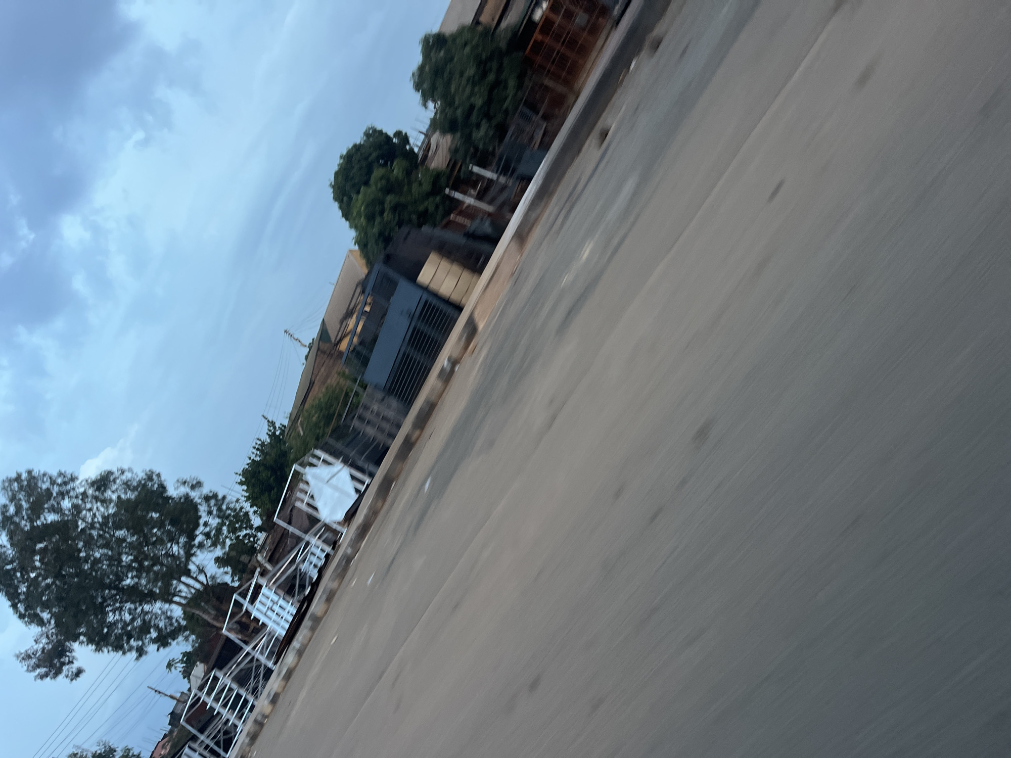 The image captures a street scene in Gura Top, Nigeria, under an overcast sky. The perspective is tilted, suggesting movement, likely from a vehicle. A paved road occupies the foreground, its surface appearing smooth and grey. Along the right side of the road, a series of metal structures, possibly scaffolding or fencing, are visible. Behind these structures, buildings and dense green trees are present. One notable feature is a large tree with dark green foliage located towards the left side of the image. The sky is filled with grey clouds, indicating a cloudy or possibly rainy day. There are no people visible in the frame.