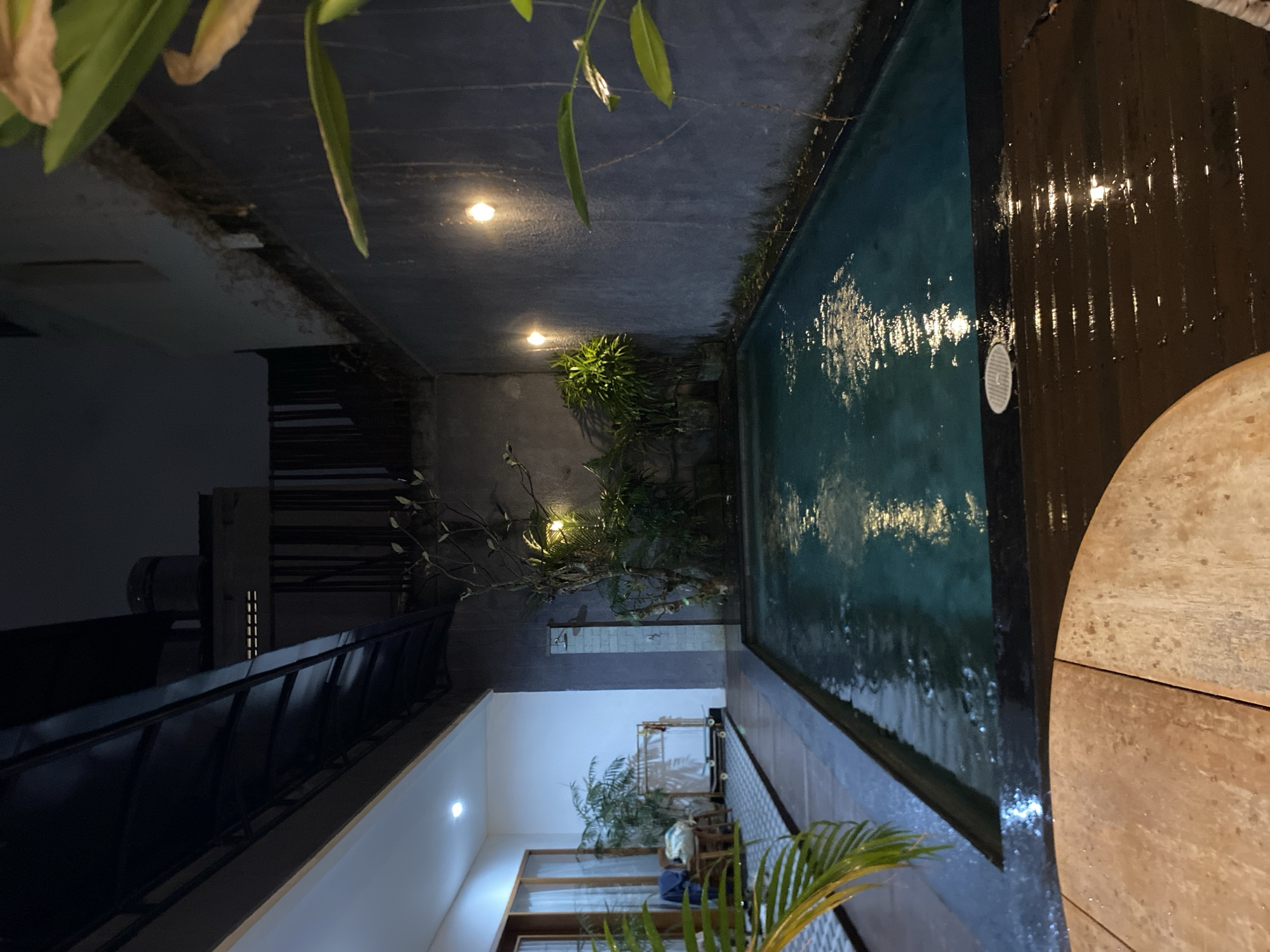 The image shows an outdoor swimming pool at night. The pool is rectangular and filled with blue water. The surrounding area is paved with dark wood planks on one side and grey concrete on the other. There are some plants near the pool, including a potted palm in the foreground. Lights are visible both in the pool area and in an adjacent building, casting reflections on the wet surfaces. The building appears to be a modern structure with dark tinted windows and a metal railing. The overall mood is tranquil and relaxing, suggesting a private residence or a resort. The time of day is clearly night, and the weather appears to be calm, as indicated by the still water in the pool and the reflections. The location context is North Kuta, Indonesia, which is known for its resorts and villas. There is no visible text in the image.