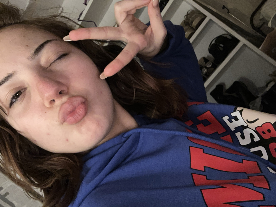 A young woman winks, pursing her lips. She holds up three fingers, painted with light pink nail polish. She wears a blue hooded sweatshirt with red lettering. Behind her is a white shelf unit, some compartments filled with dark shoes or accessories. The lighting is soft, creating a casual indoor scene.