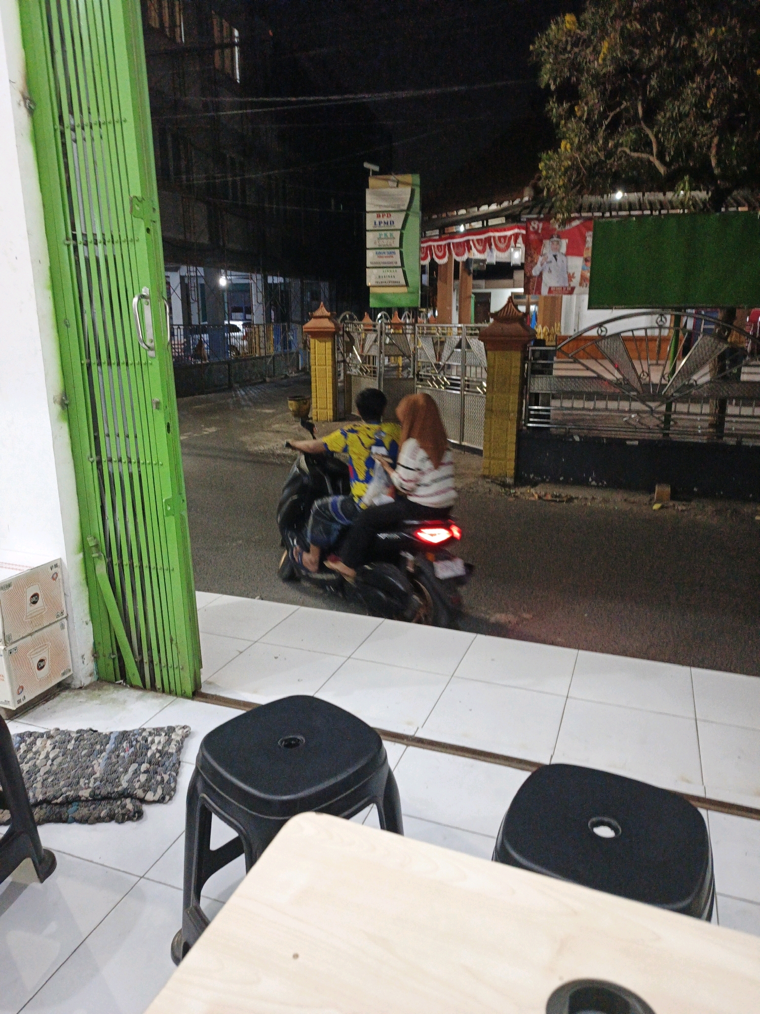 In the city of Ketegan, Indonesia, a nighttime scene unfolds outside a building with a bright green collapsible gate. A man and a woman ride a black motorcycle with its taillight illuminated. The man, wearing a vibrant yellow and blue patterned shirt and dark pants, drives the motorcycle. The woman, seated behind him, wears a white and red striped long-sleeved shirt and a rust-colored hijab. They are seen from behind, as they ride away from the viewer.
In the foreground, a black plastic stool is visible on a tiled floor. To the left, a mat made of what appears to be small stones and fabric lies on the floor. Boxes are stacked against the building wall, near the green gate.
The background reveals a street scene at night. Buildings with various signs are illuminated. One sign, partially visible, has green and white elements. Another features red and white decorations. A large tree with yellow flowers is prominent in the upper right of the frame. A decorative metal fence separates the building from the street.