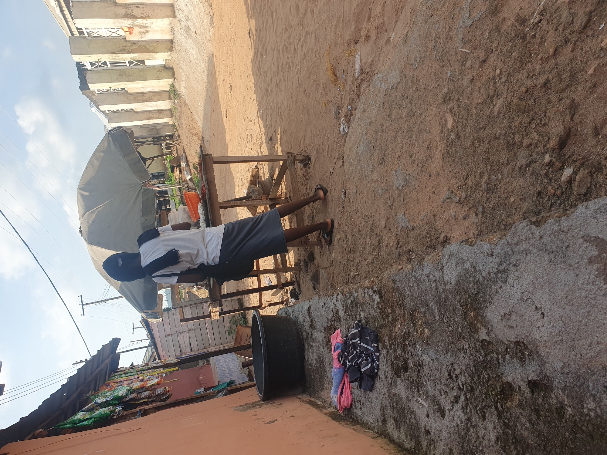 A young male, wearing a white t-shirt and grey shorts, stands near a market stall in Lokoja, Nigeria. He is partially obscured by a large, grey umbrella that shades the stall. The market appears to be set up outdoors, with a sandy ground and a concrete barrier in the foreground. Behind the stall, there are some structures with corrugated iron roofs.
In front of the young man, to his right, is a large, black, circular container, possibly for washing or storage. To the right of the container, a pile of folded clothes is visible on the concrete. The clothes are a mix of colors and patterns, including pink, blue, black, and white stripes.
The background shows a building with multiple levels and balconies, suggesting an urban or semi-urban environment. Power lines are also visible against a bright, partly cloudy sky, indicating daytime. The overall impression is of a typical market scene in a warm climate. The lighting suggests it is daytime, likely morning or afternoon, with the sun casting shadows. There are no discernible emotions visible on the subject's face due to the angle and partial obstruction.
