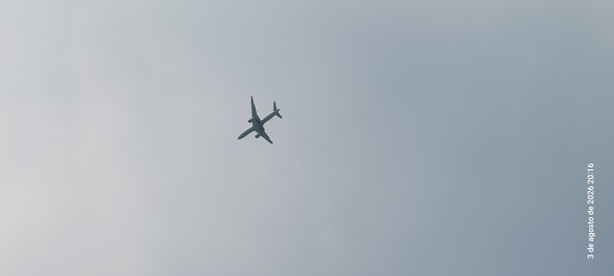 A commercial airplane is shown from below, in flight against a pale, overcast sky. The date and time stamp "3 de agosto de 2026 20:16" is visible in the lower left corner of the image. There is no indication of the city of Castelldefels, Spain within the image itself.