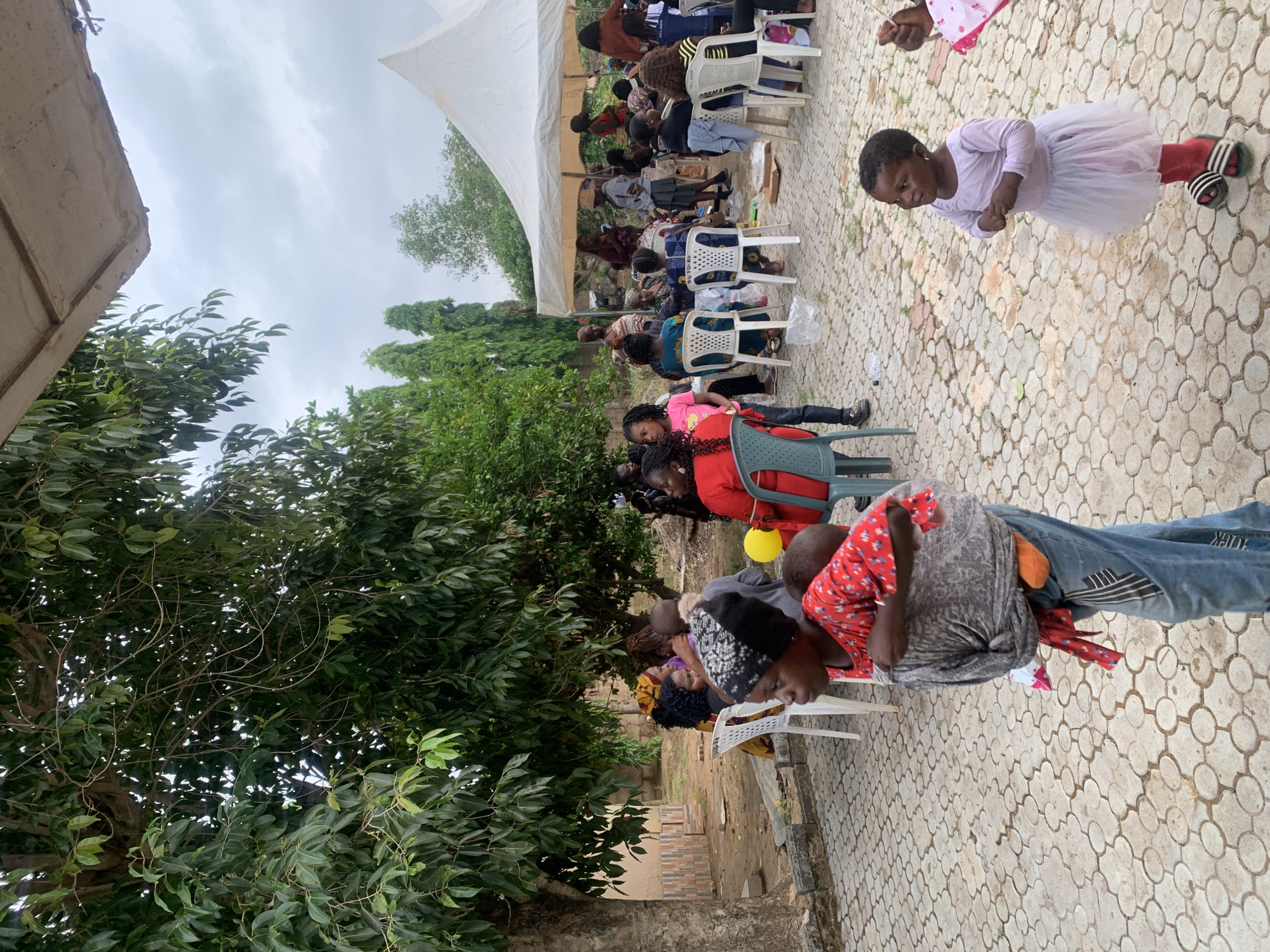 A gathering of people is seated in white chairs beneath a white canopy in an outdoor setting. A young girl stands in the foreground wearing a light-colored dress and dark shoes. Individuals are seated in rows, and some appear to be watching something. The location is identified as Gura Top, Nigeria. The ground is paved with a circular patterned design. Green trees surround the area. The sky is overcast.
