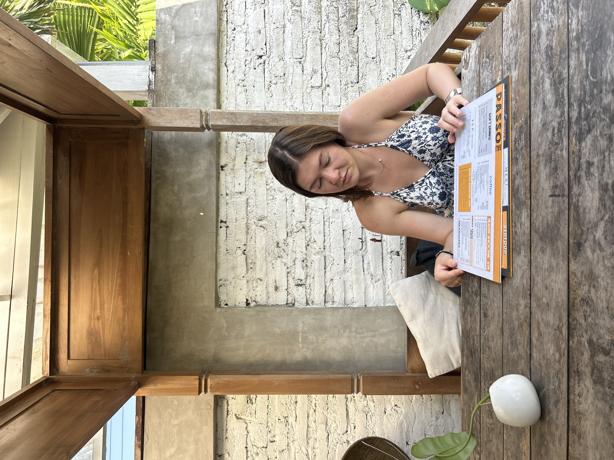 A young woman is seated at a wooden table outdoors, looking down at a menu. She is wearing a white and blue floral print bikini top and a delicate necklace. The table is made of weathered wood planks. To the left of the woman, on the table, is a small white vase containing a green leaf. Behind the woman is a white brick wall with a textured gray plaster section. A wooden structure with carved pillars frames the scene. To the right of the woman, the environment suggests a tropical location with palm fronds visible in the background. The woman appears to be in the city of Mengwi, Indonesia, likely at a restaurant or cafe.