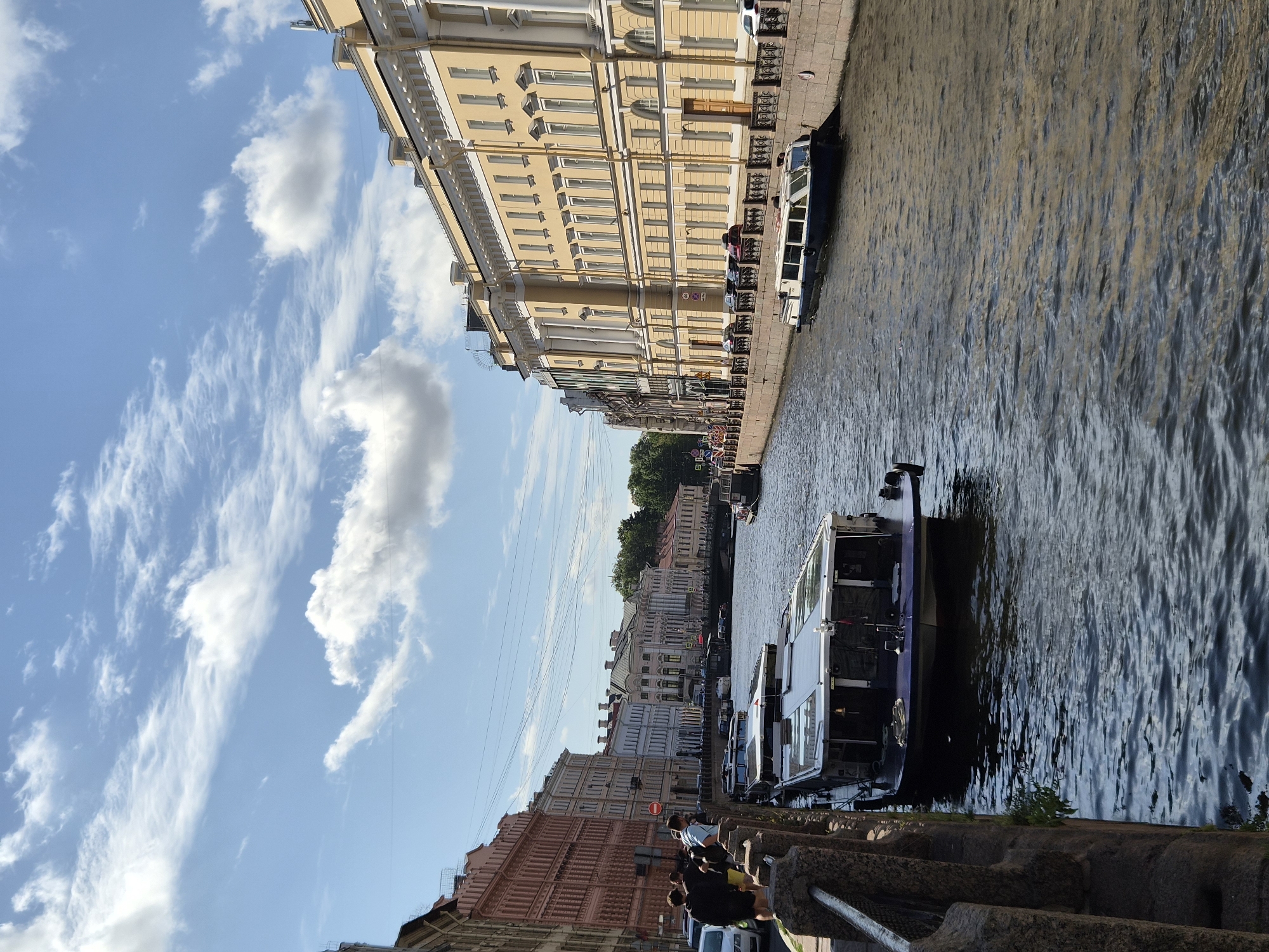A sunny daytime scene depicts a wide canal in a city, bordered by ornate buildings on both sides. Several boats are docked or cruising on the water. On the left side of the canal, a row of multi-story buildings with pastel-colored facades lines the waterfront. People are visible walking along the sidewalk and standing near the edge of the canal. Several cars are parked along the street in front of the buildings.
On the right side of the canal, a large, pale yellow building with classical architectural elements dominates the view. It features numerous windows and decorative columns. A black metal fence runs along the edge of the canal, separating the sidewalk from the water.
The canal itself is filled with dark, rippling water, reflecting the sky and buildings. Two prominent boats are in the foreground. The first, on the left, is a long, dark-colored vessel with a glass-enclosed upper deck. The second, further down the canal on the right, is a blue and white boat with large windows. Other smaller boats and barges are visible further in the distance, as well as a bridge spanning the canal. The sky above is blue with scattered white, fluffy clouds, some of which are wispy. Power lines crisscross the upper portion of the image, strung between poles. The overall impression is of a bustling urban waterway on a pleasant day.