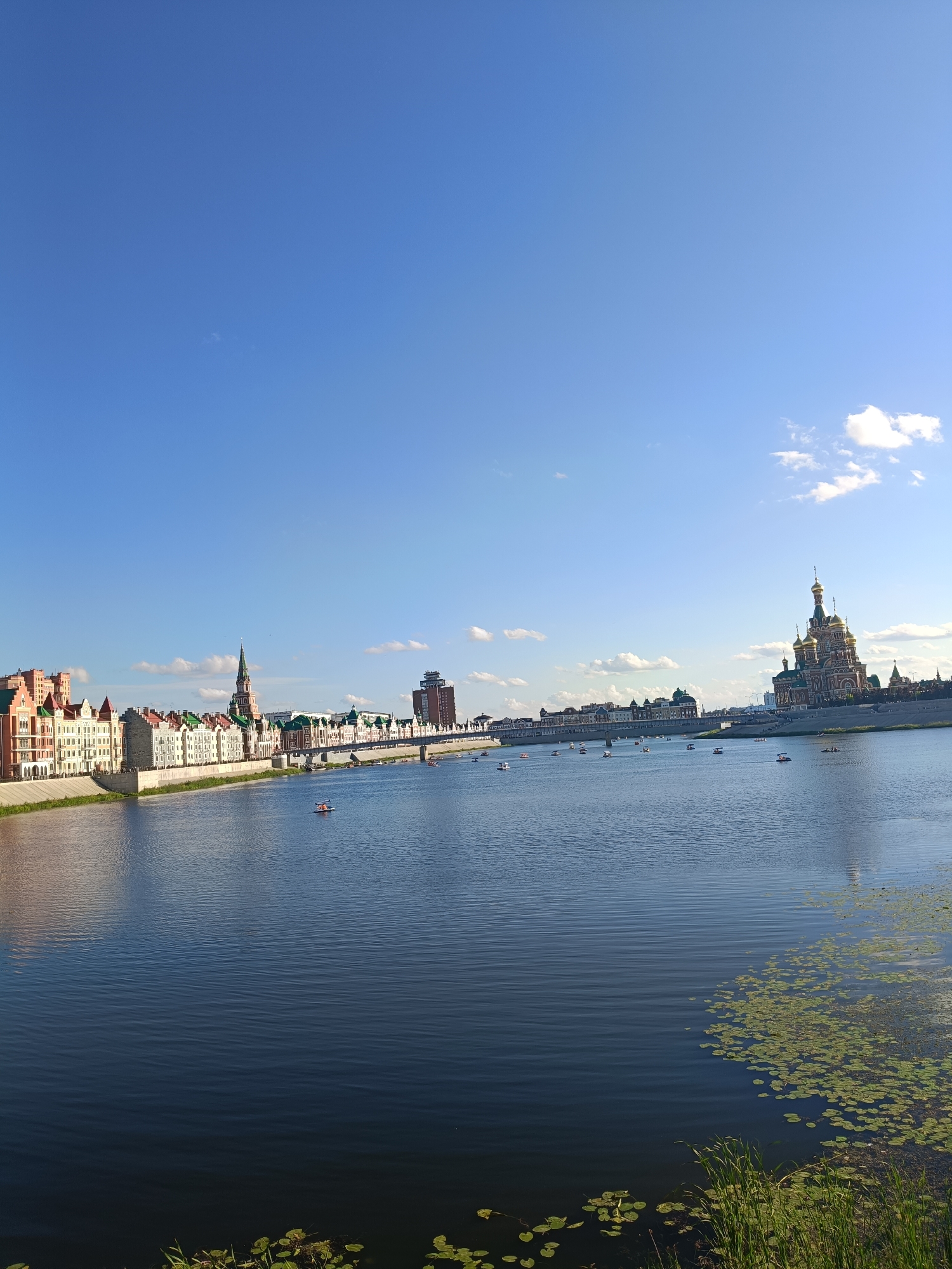 The image shows a panoramic view of the Yoshkar-Ola embankment in Russia. The scene is dominated by a wide, calm river under a clear blue sky with scattered white clouds. On the left bank of the river, a row of colorful, European-style buildings with red and green roofs lines the embankment. A prominent, tall, pointed spire of a building stands out among them. Further down the embankment, a long bridge spans the river, with several small boats and paddle boats visible on the water, some with people in them. On the right bank, a large, ornate cathedral with golden domes is a focal point, with other buildings and structures visible behind it. The foreground of the image, particularly on the right side, shows the river's edge with lily pads and tall green grass. The overall impression is one of a sunny day in a picturesque city.