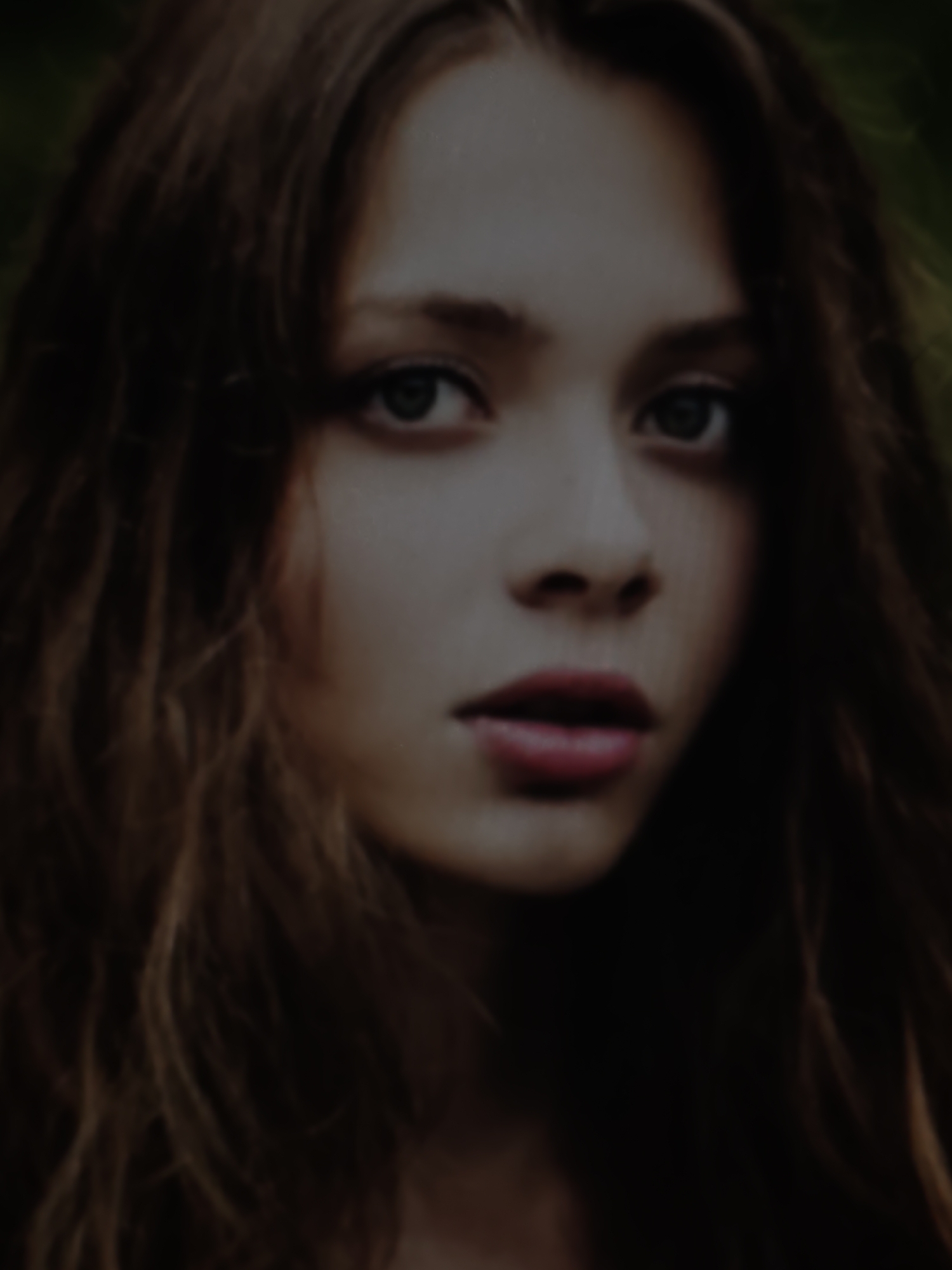 The image is a close-up, slightly blurred portrait of a young woman with long, dark, wavy hair. Her eyes are looking directly at the camera, and her lips are slightly parted. The lighting is dim, creating a moody atmosphere. The background appears to be a natural setting, possibly outdoors with foliage, but it is out of focus. There is no visible indication of the city of Kurganinsk, Russia, or any specific urban environment.