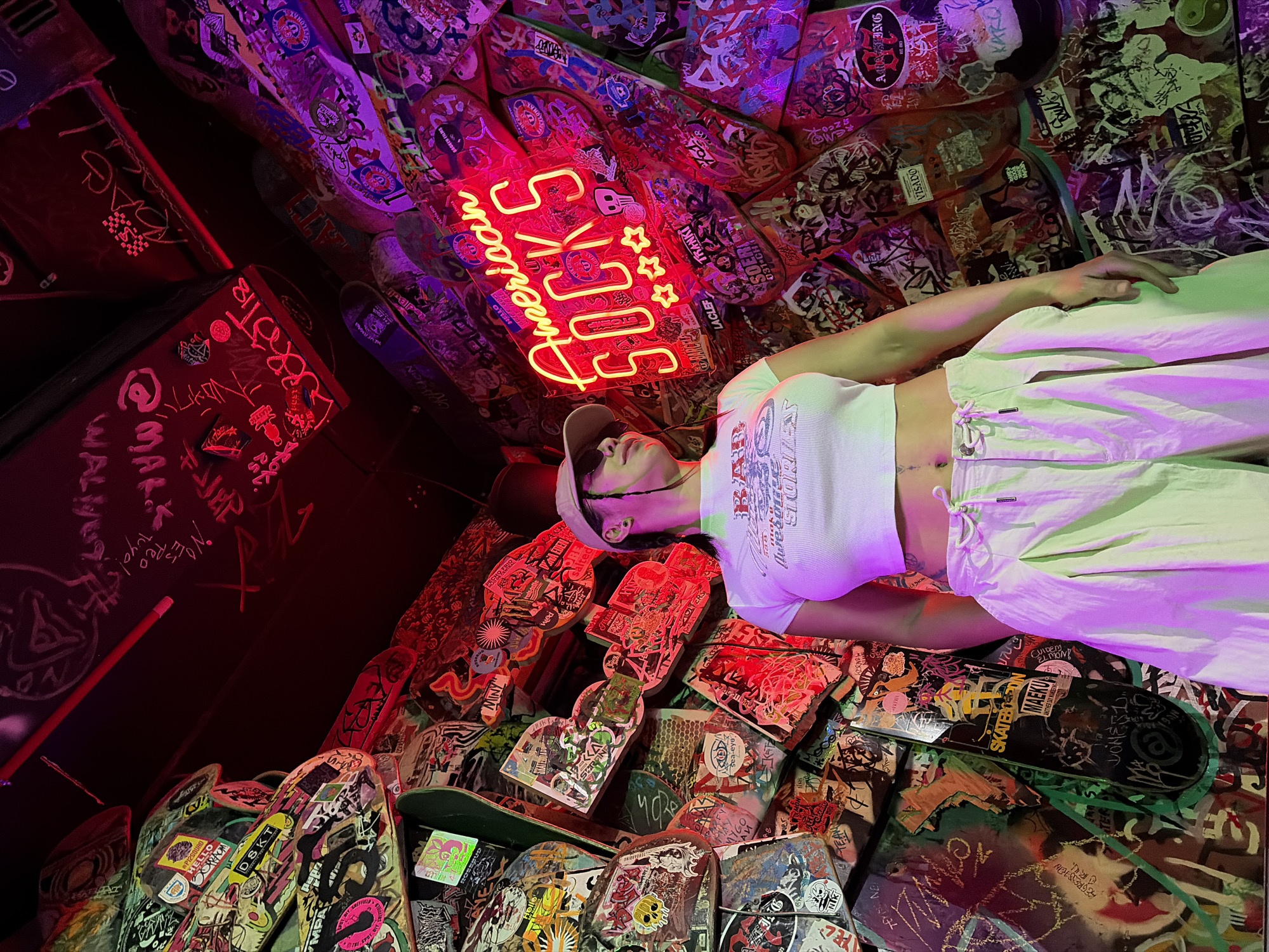 This vibrant indoor scene, set in Barcelona, Spain, features a person lying amidst a densely decorated environment predominantly composed of skateboard decks. The subject, a female-presenting individual, is positioned centrally, wearing a white crop top, light-colored baggy pants, sunglasses, and a baseball cap. She is looking upwards, or slightly to her right, as if observing the overhead decor.
The activity appears to be a posed photograph, capturing the individual within this unique, edgy space. The room is dimly lit, dominated by the glow of a bright neon sign that reads "American SOCKS" accompanied by three stars, casting a strong pink and red hue over the area. Additional purple and green ambient lights further enhance the colorful, dynamic atmosphere.
Notable details include the overwhelming number of skateboard decks covering the walls and possibly the ceiling, creating a mosaic of skate culture. These decks are adorned with countless stickers, graffiti, and custom artwork, reflecting a rebellious and artistic aesthetic. Visible text on the decks and walls includes brand names, slogans, and tags such as "DSKT," "MAEKU," "SKATEBOARD," "SOERI," "LACLEF," "TRANKI," "BROKEN FILMS," "VISADO," and "Red Bull." Other discernible graffiti includes "@MAL WAL.K" and "Tuyo!". The overall impression is one of a contemporary, urban, and creatively charged environment, likely a skate shop, bar, or art installation, captured during the nighttime or in a windowless space.