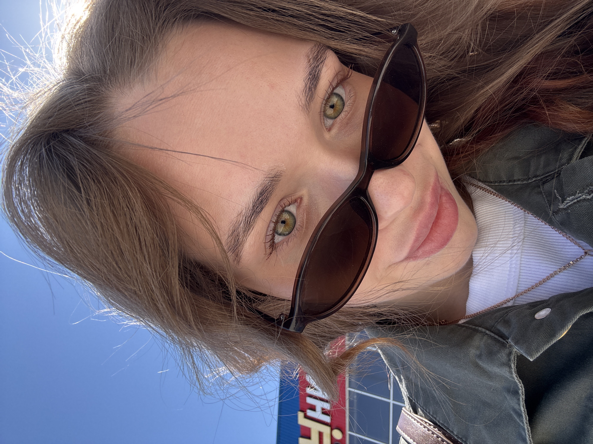 A woman takes a selfie outdoors in Chaikovsky, Russia. The woman has long, light-brown hair and green eyes, wearing sunglasses, a white shirt, and an olive green jacket. She is facing the camera at an angle. In the background, a blue sky is visible, as well as a section of a blue and red building.